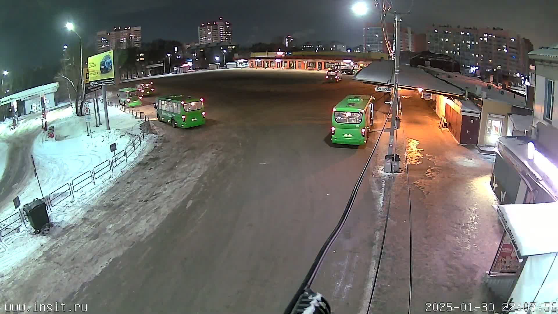 A nighttime view of a bus station with several green buses parked, surrounded by snow-covered ground and buildings under streetlights.