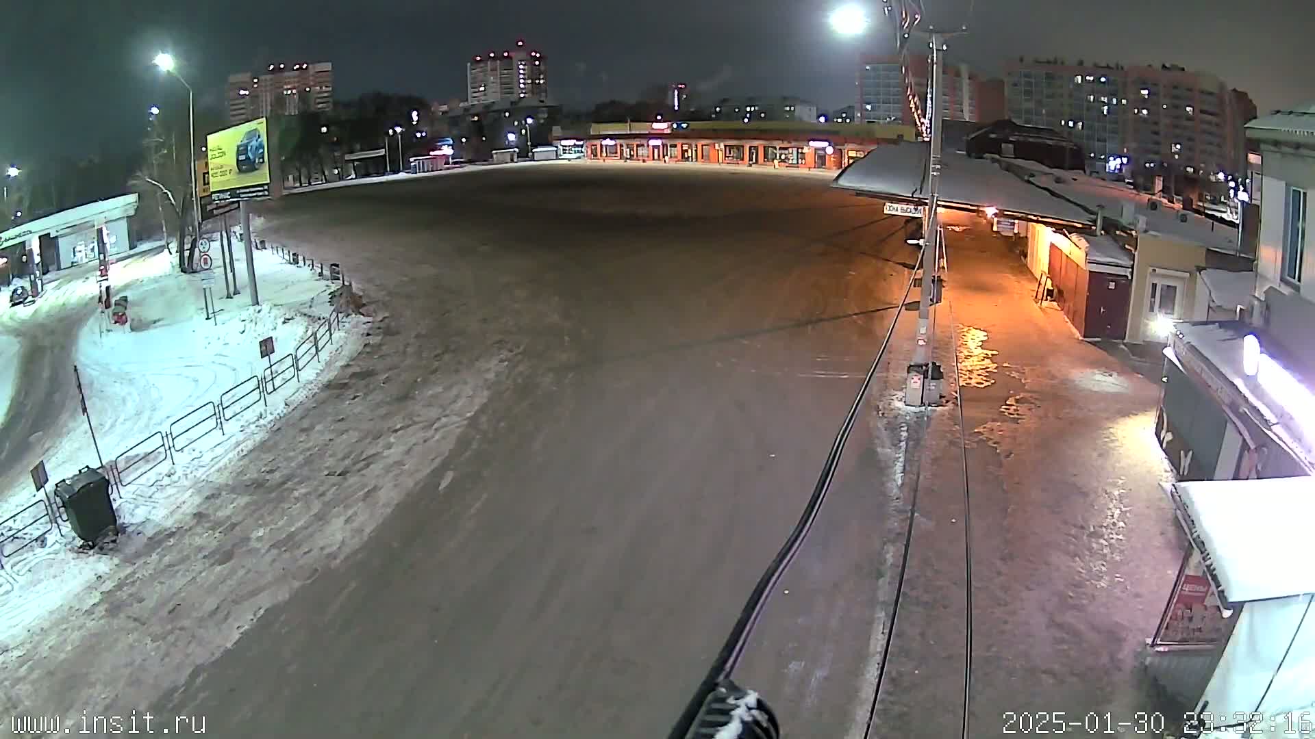 A snow-covered, mostly empty plaza at night is seen from a high vantage point, with buildings and streetlights visible in the background.