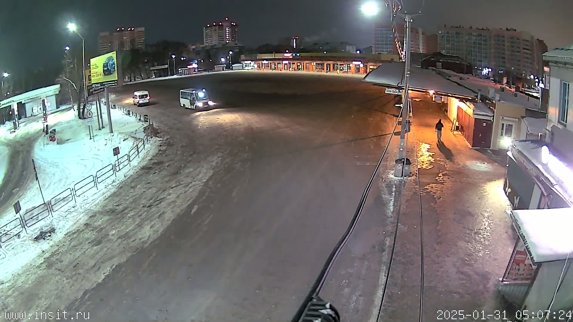 A nighttime, snowy scene shows a mostly empty parking lot with a few vehicles and a pedestrian, under streetlights.
