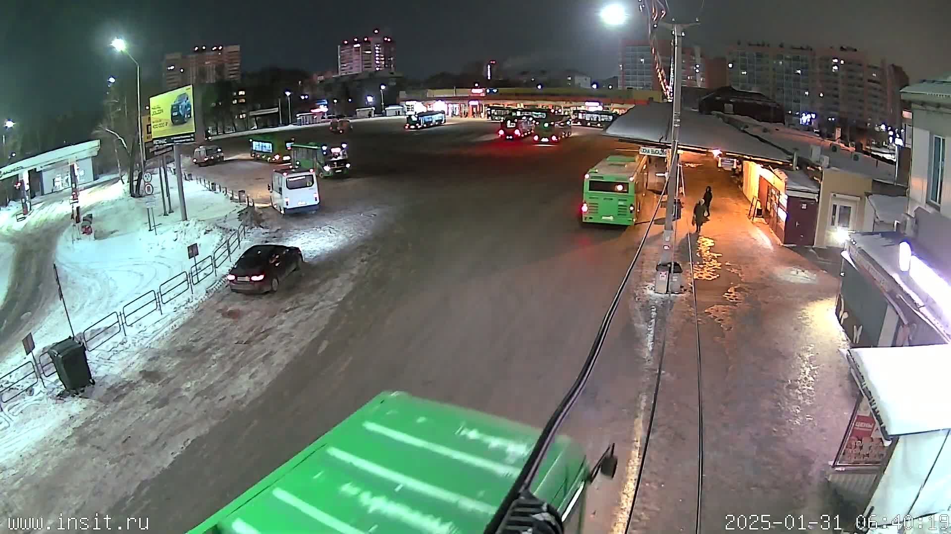 A nighttime scene of a snowy bus station with several buses parked and a few cars, with pedestrians walking nearby.