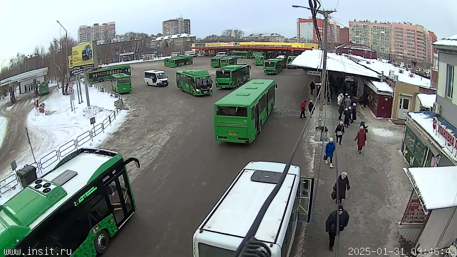 A bus depot with numerous green buses and a few pedestrians, covered in snow, under an overcast sky.
