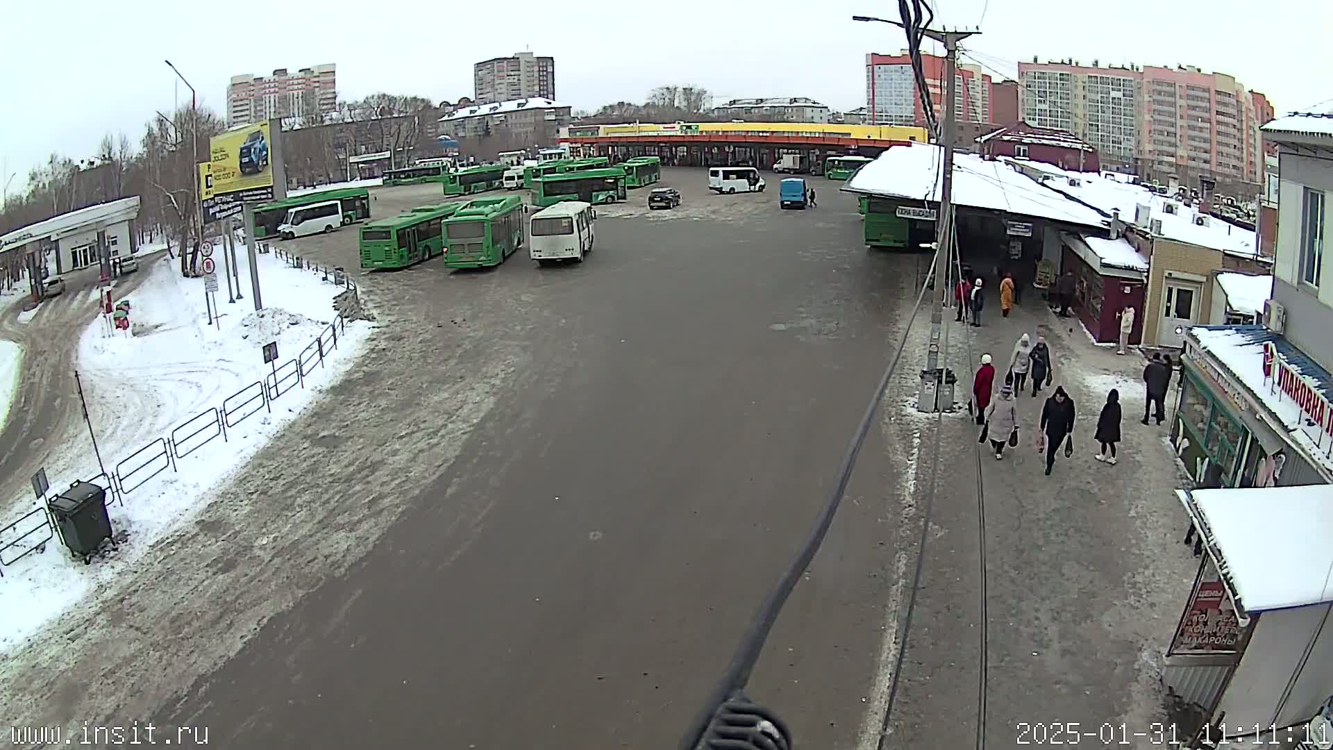 A bus station with numerous green buses parked, some cars, and several people walking around in snowy winter conditions.