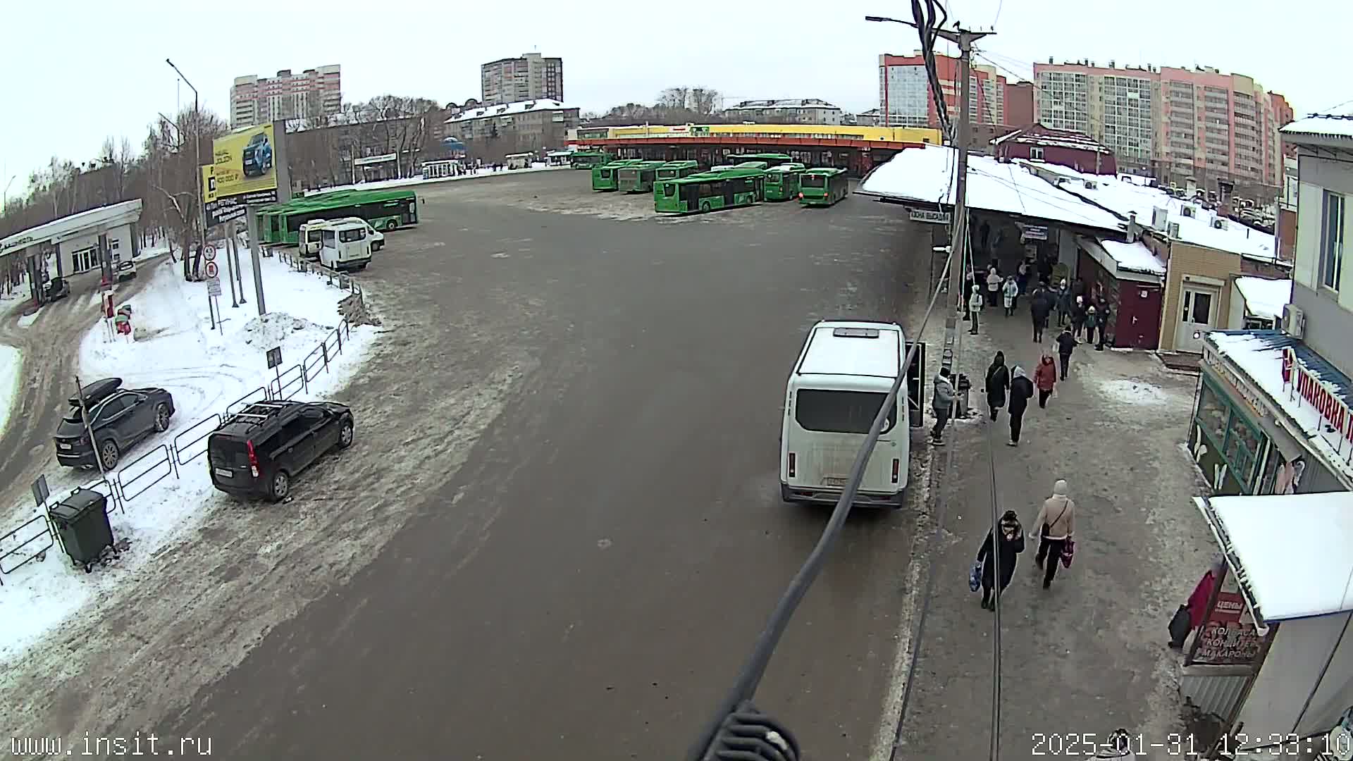 A snowy bus station with several green buses parked, a few cars, and people walking around.