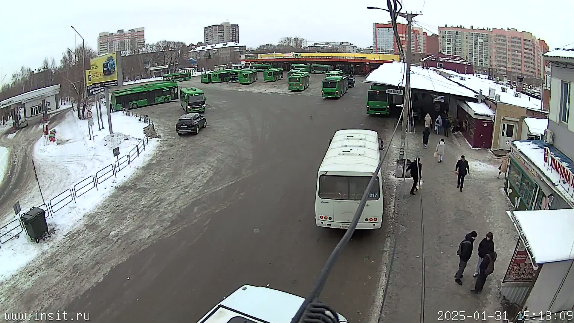 A bus station with numerous green and white buses, several cars, and pedestrians, is partially covered in snow on an overcast day.
