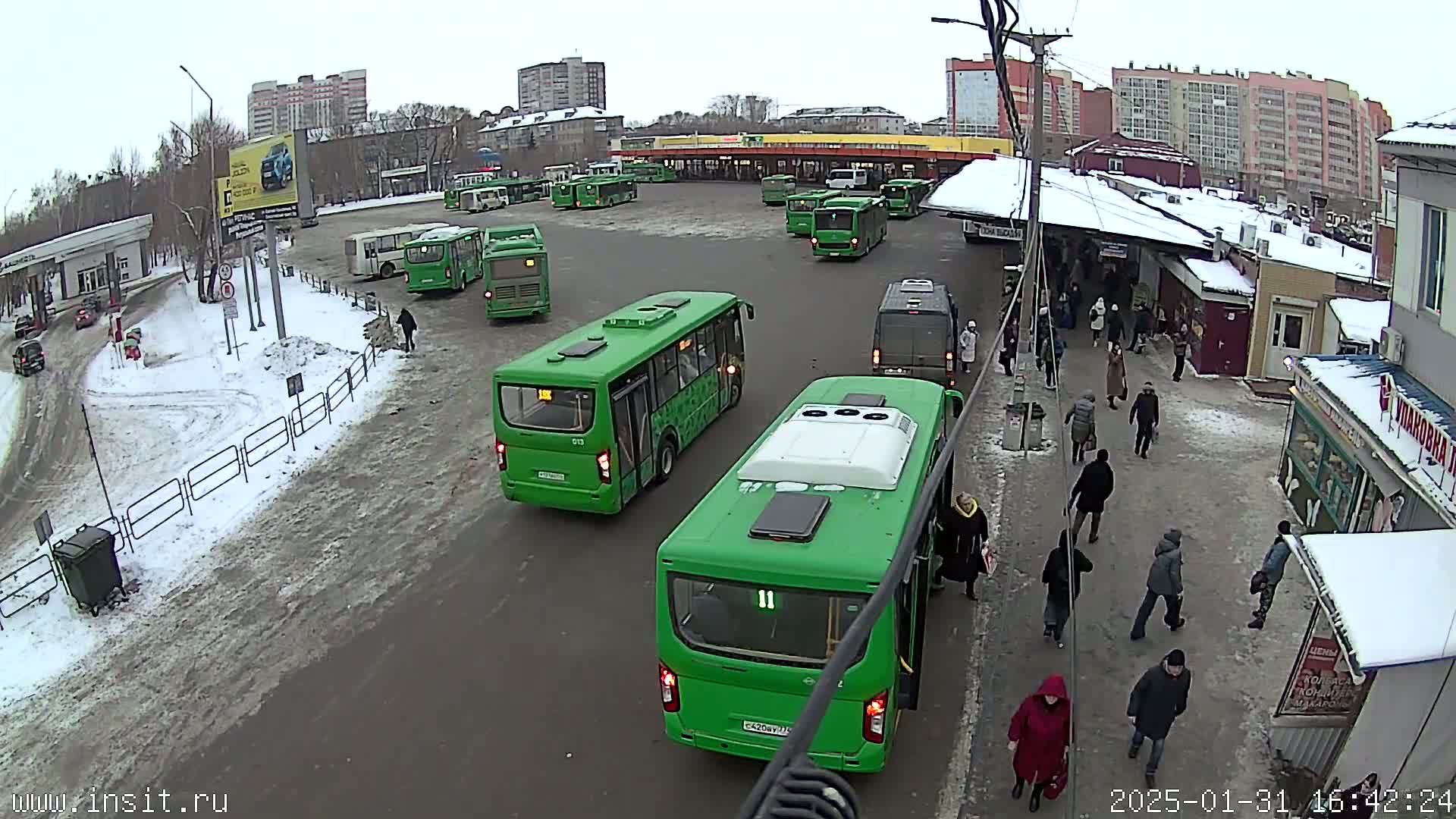 A bus station with numerous green buses and several people walking around in snowy conditions.