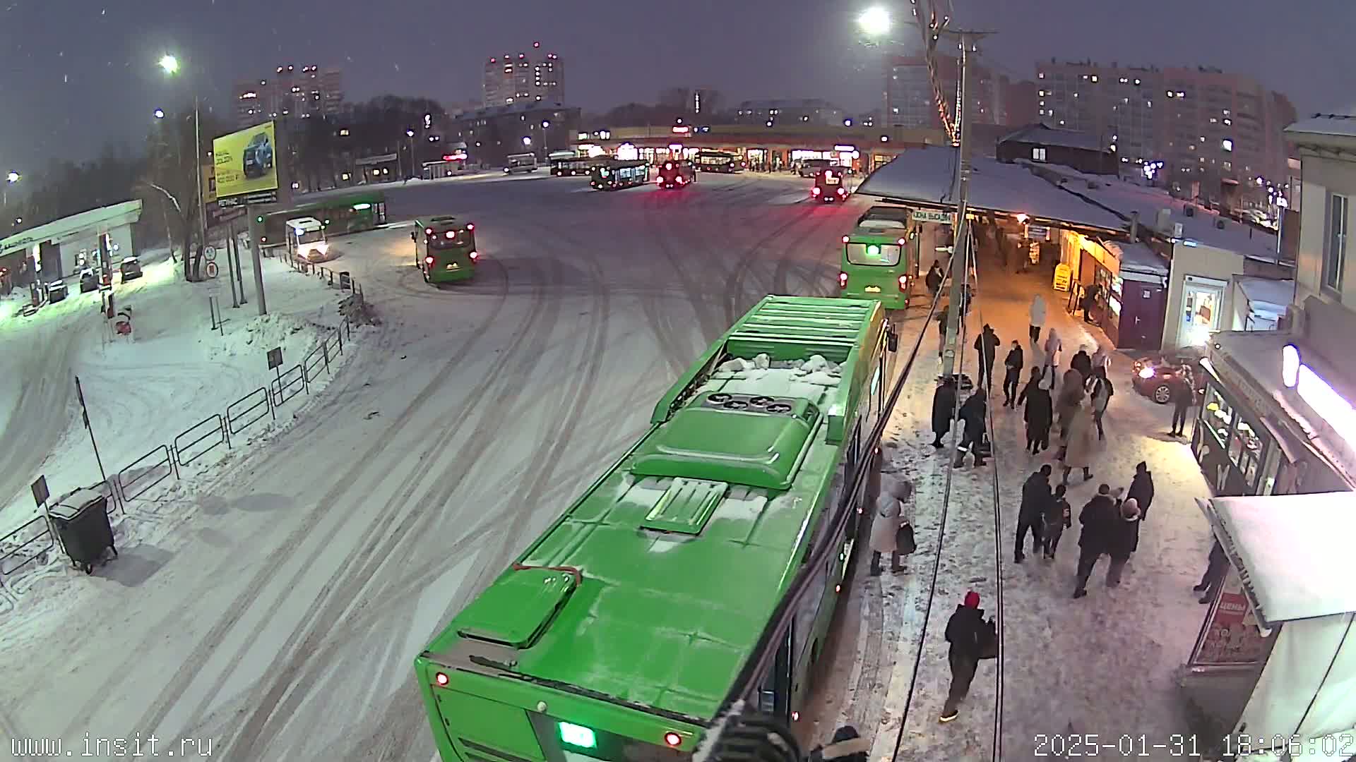 A snow-covered bus depot at night with several green buses and people waiting on the snowy ground.