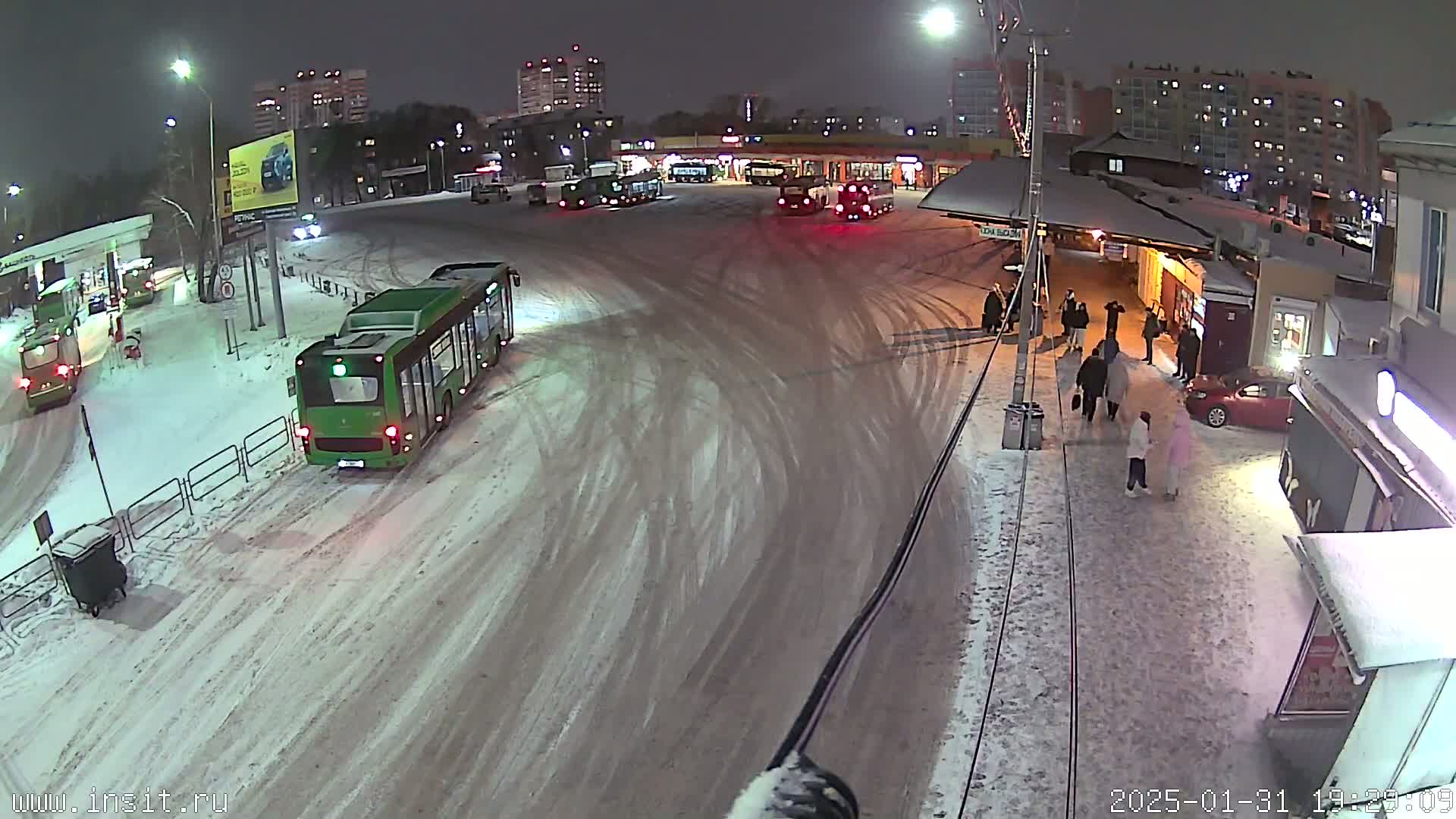 A snow-covered bus station at night, with several buses parked and a few people walking around.