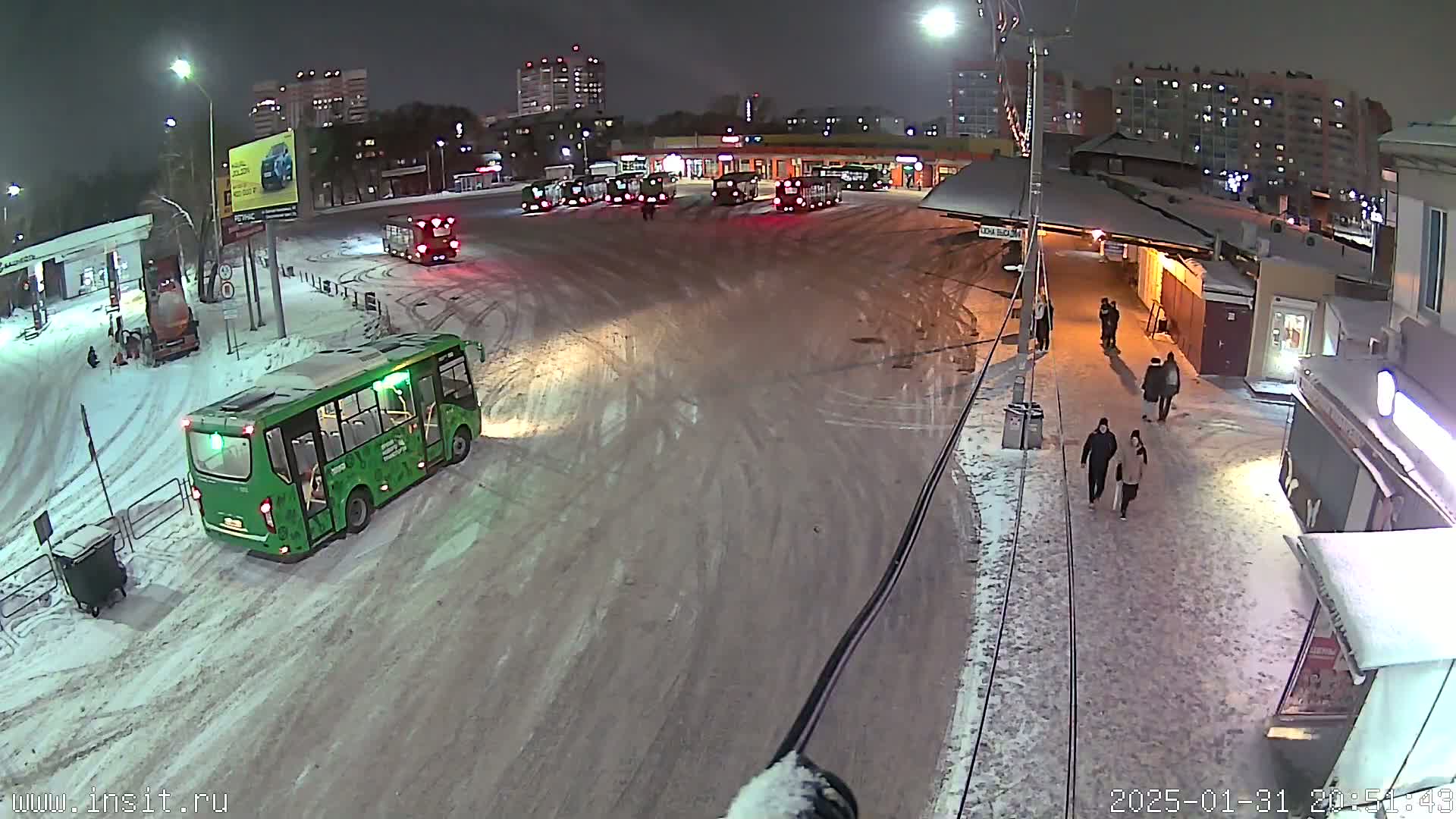 A snow-covered bus depot at night, with several buses parked and a few pedestrians walking along a snow-covered street.