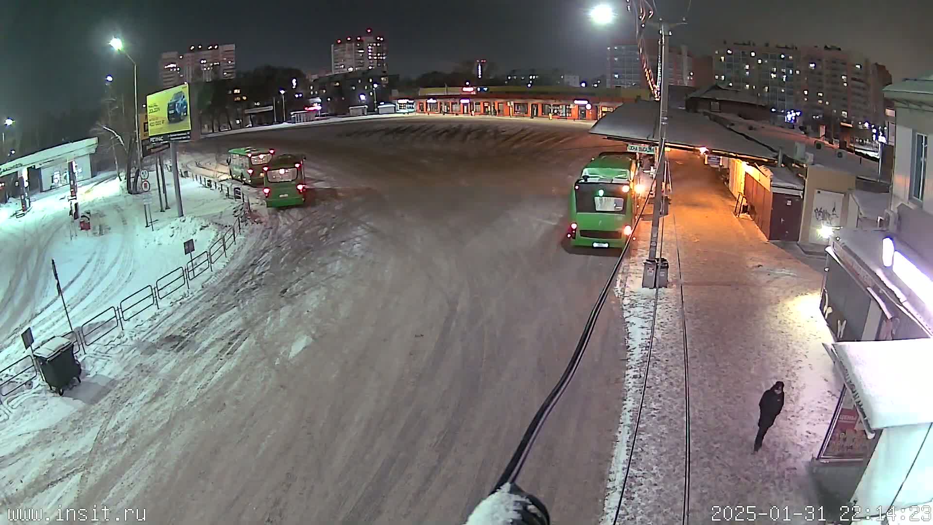 A snow-covered nighttime scene shows a bus station with several buses and a lone pedestrian walking.