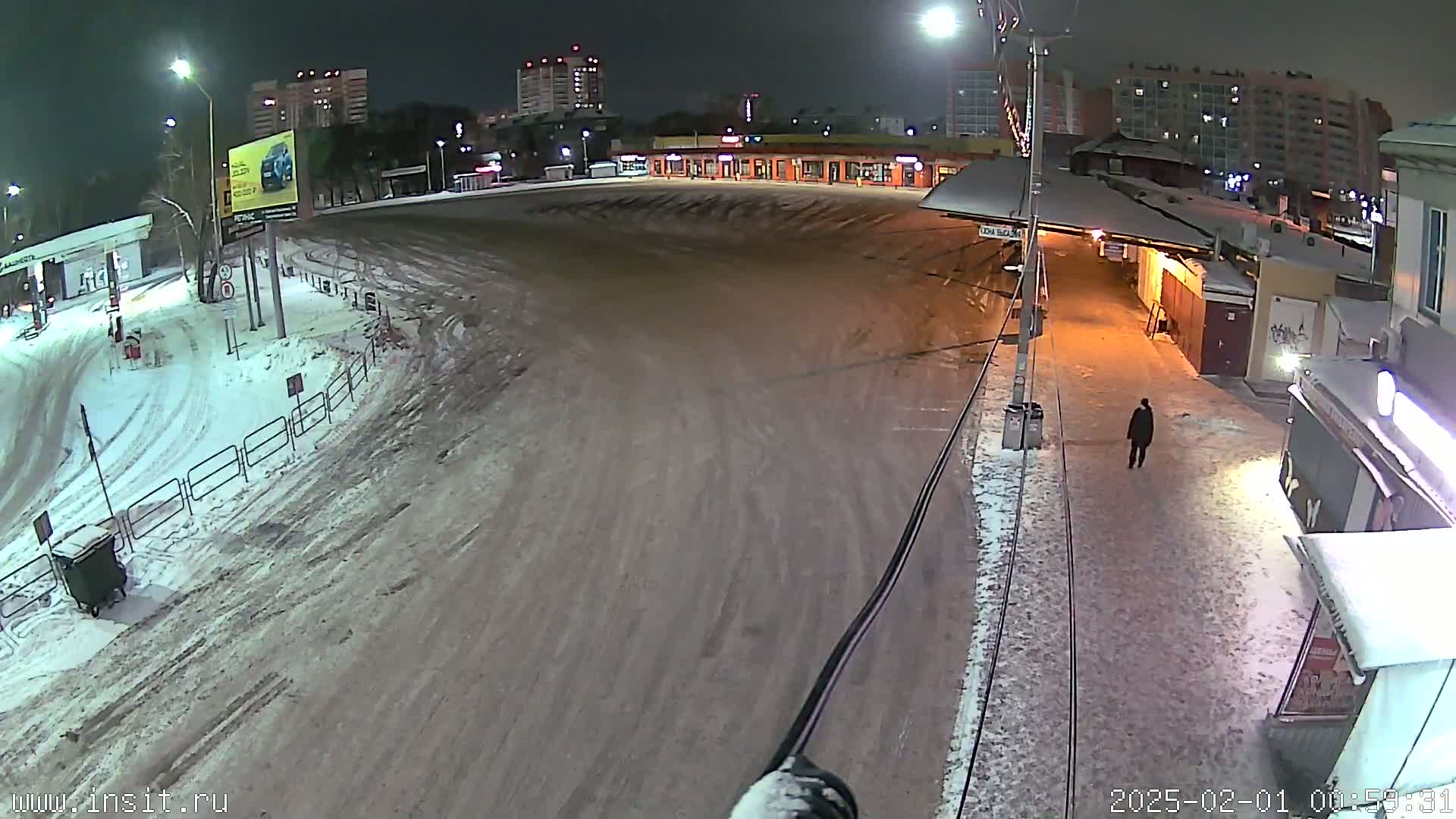 A snow-covered parking lot and roadway are illuminated at night, with a lone person walking near some buildings.