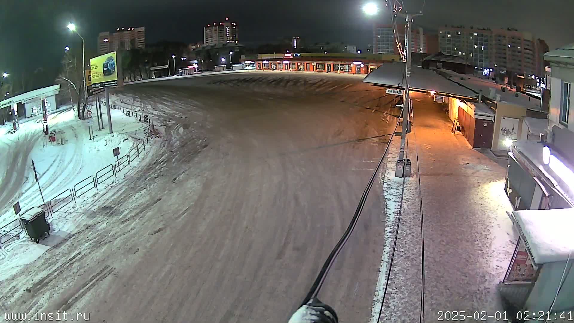 A snow-covered, empty plaza at night, with buildings and streetlights in the background.