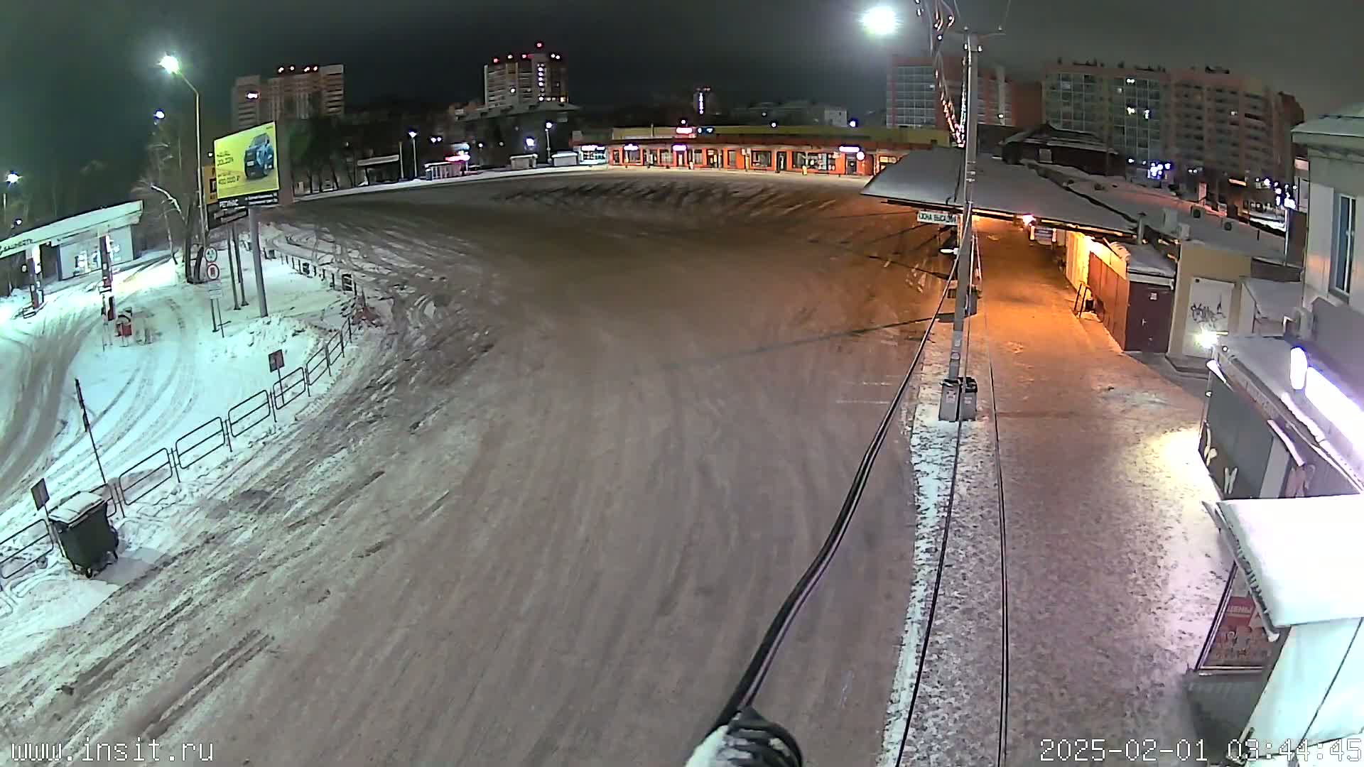A snow-covered empty plaza at night, with buildings and streetlights visible in the background.