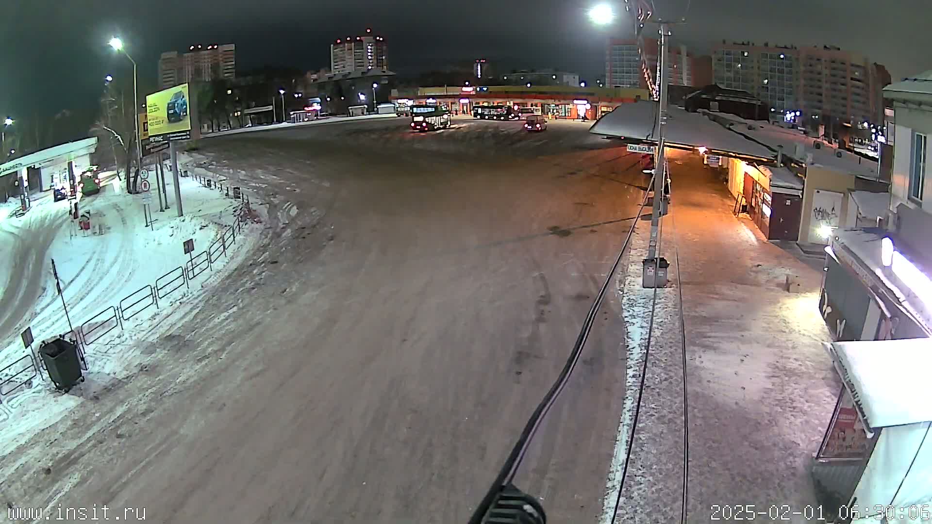 A nighttime, snow-covered bus stop with several buses parked and a few cars visible in the distance under partly cloudy skies.