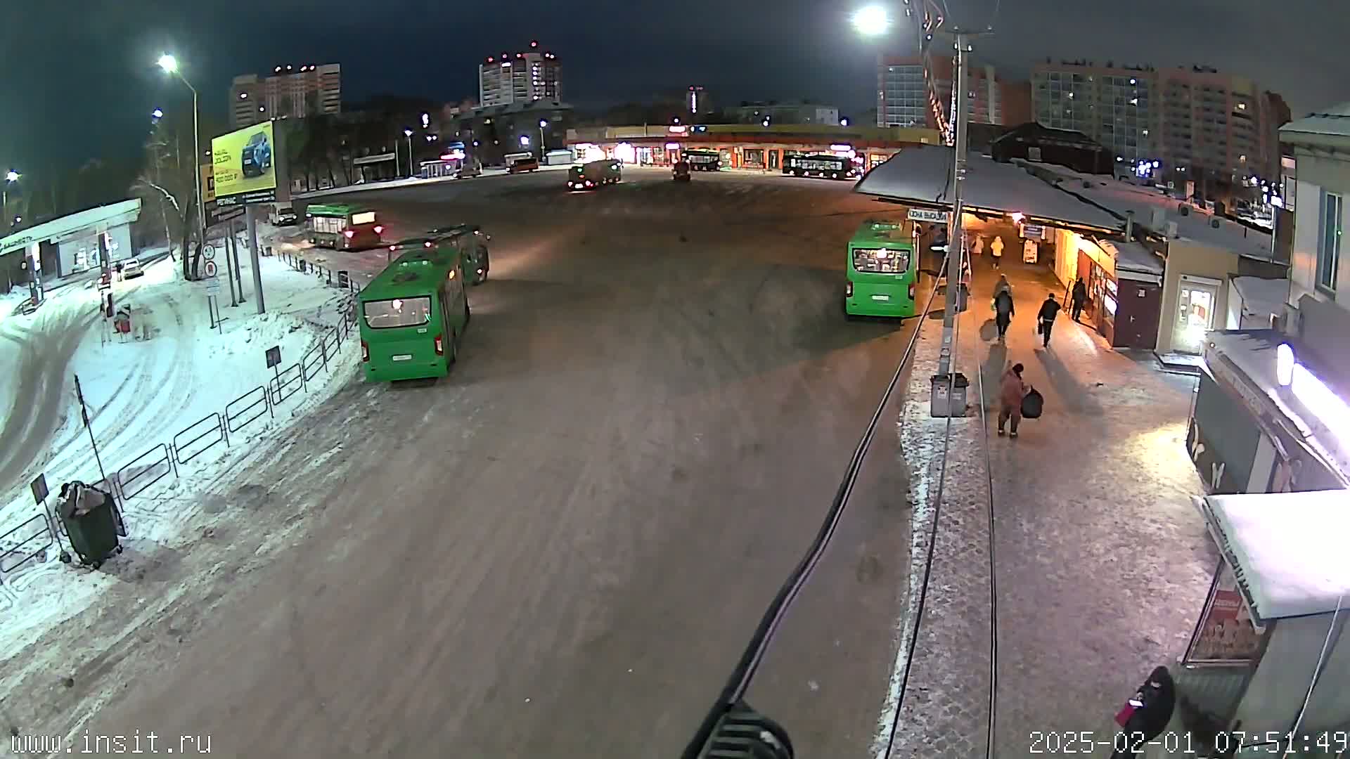 A nighttime view of a snowy bus station with several buses and pedestrians, under streetlights.