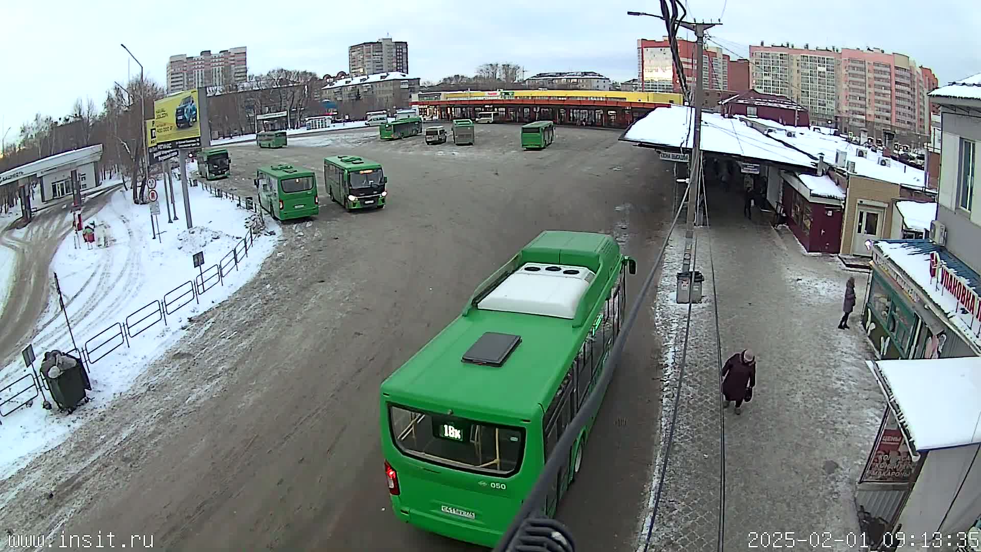 A bus depot with several green buses parked in a snowy lot, and a few pedestrians walking nearby.