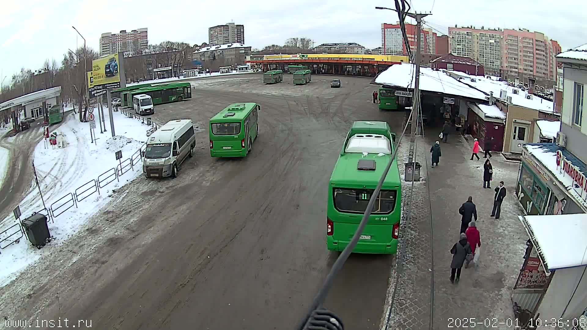 A bus depot with several green buses and a few other vehicles, in a snowy area with overcast skies.