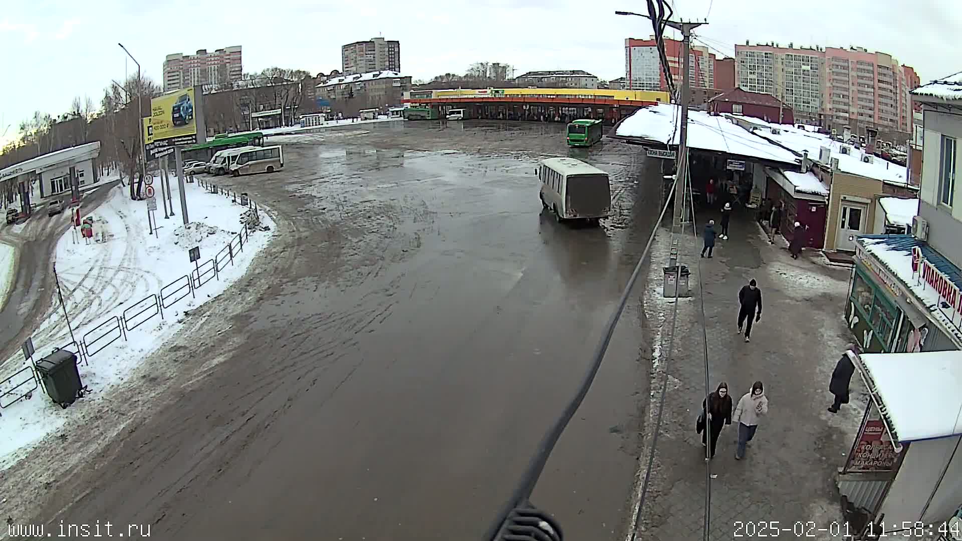 A muddy bus station with several buses and people walking around on a partly snowy, overcast day.