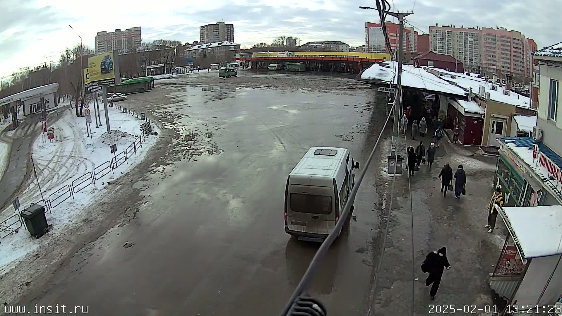 A muddy bus station with several people walking around, a white van, and buses parked, under overcast skies with patches of snow and ice on the ground.