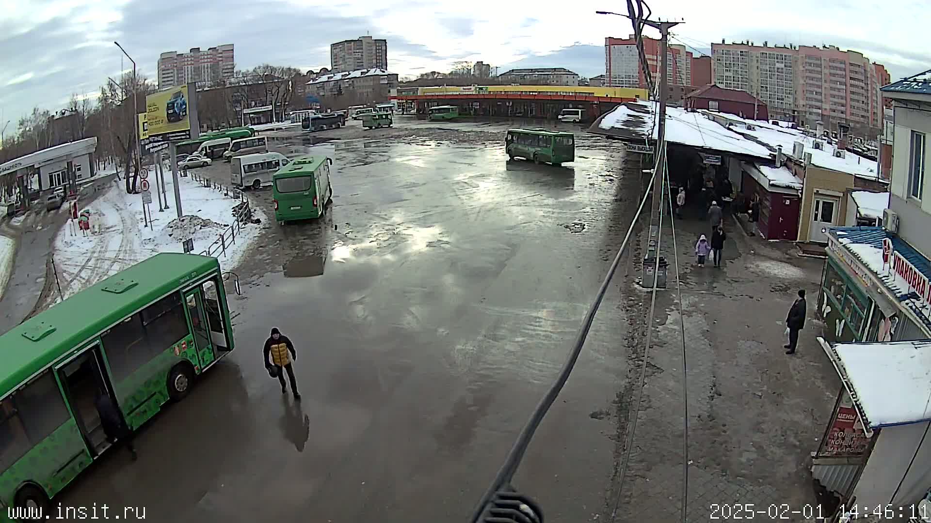 A muddy bus depot with several green buses, some cars, and a few pedestrians, under an overcast sky.