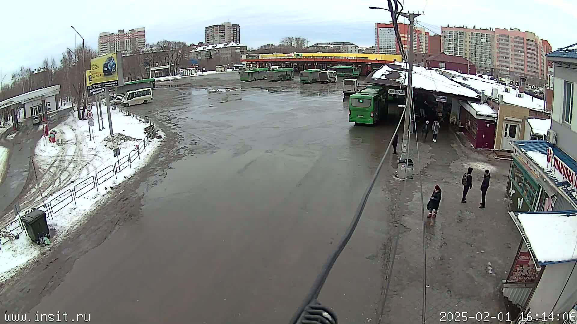 A muddy bus station with several buses parked, some people walking around, and a light snow cover on the ground in the overcast weather.