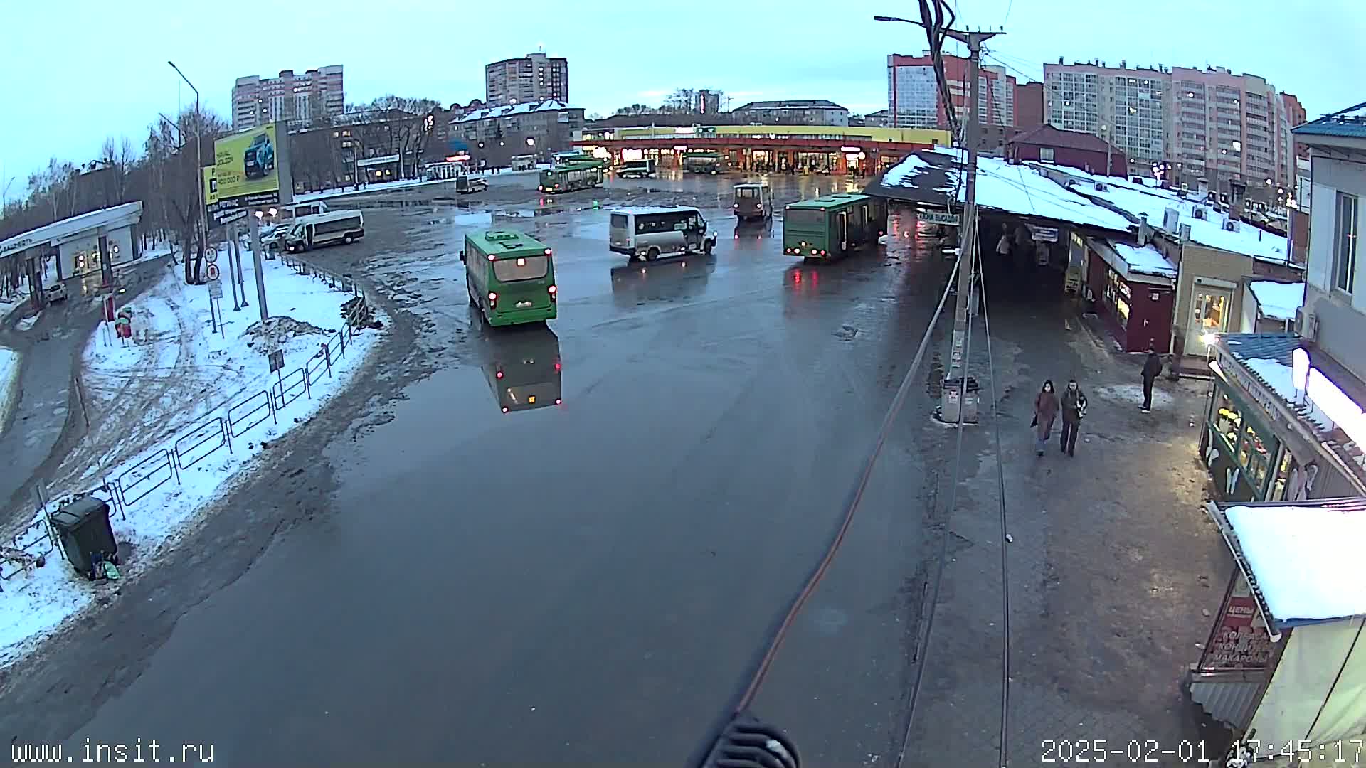 A flooded bus stop area with several buses and vans, surrounded by apartment buildings under a gloomy sky with snow on the ground.