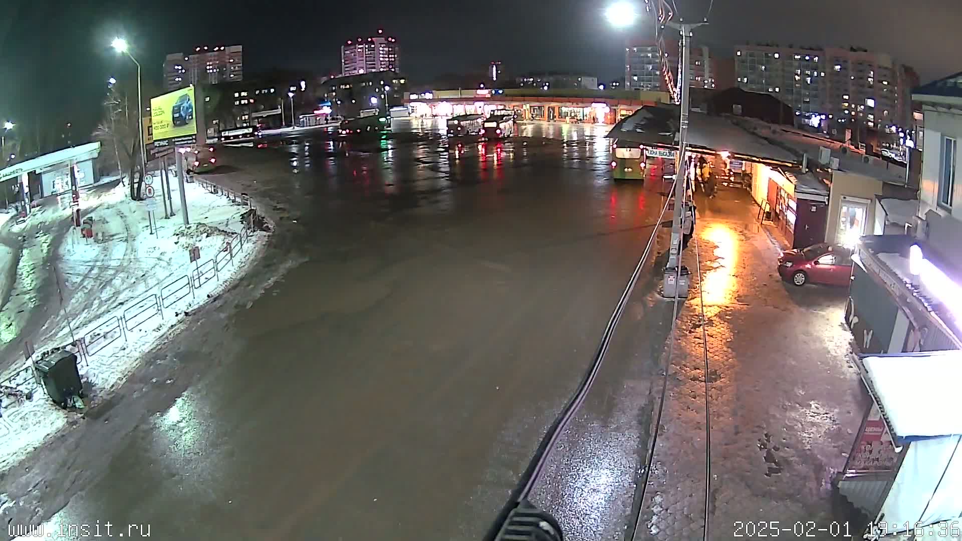 A wet, nighttime scene shows a bus station with several buses and cars parked, surrounded by buildings and patches of snow.