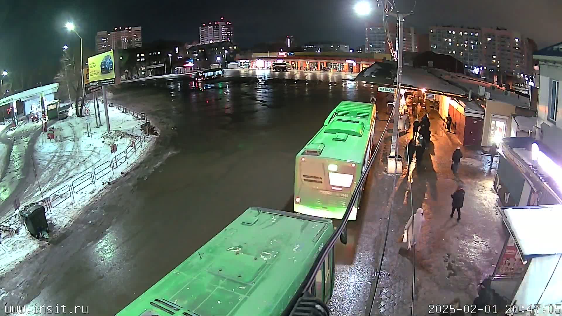 Two green buses are parked at a night bus stop in a wet, snowy area, with several pedestrians walking nearby and buildings in the background.