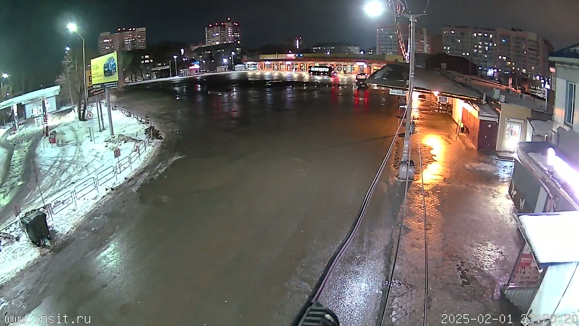 A nighttime, wet scene shows a flooded plaza area in front of buildings, with patches of snow and ice visible.