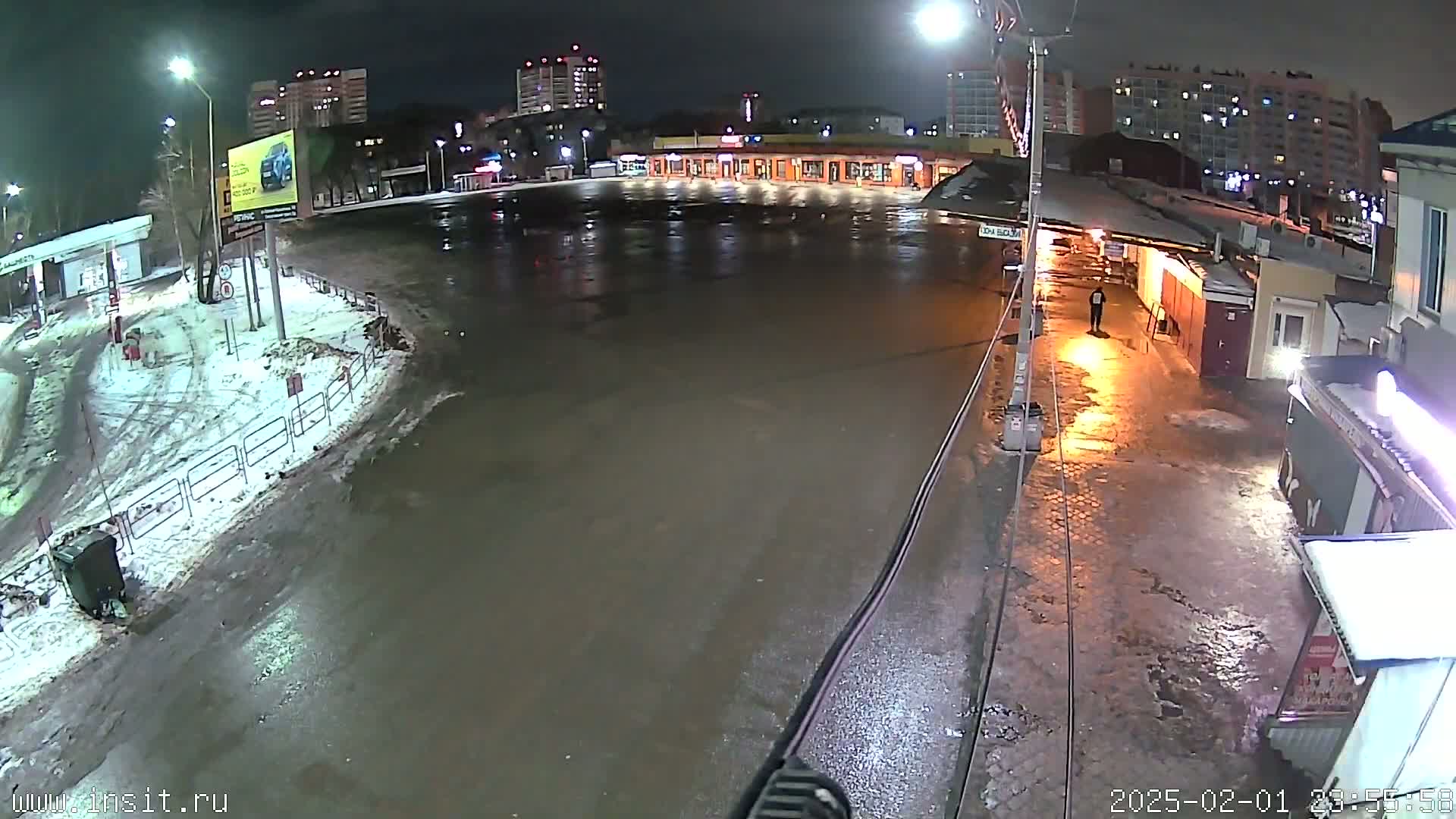 A wet, nighttime scene shows a mostly empty paved area in front of several buildings, with some snow and ice visible along the edges.