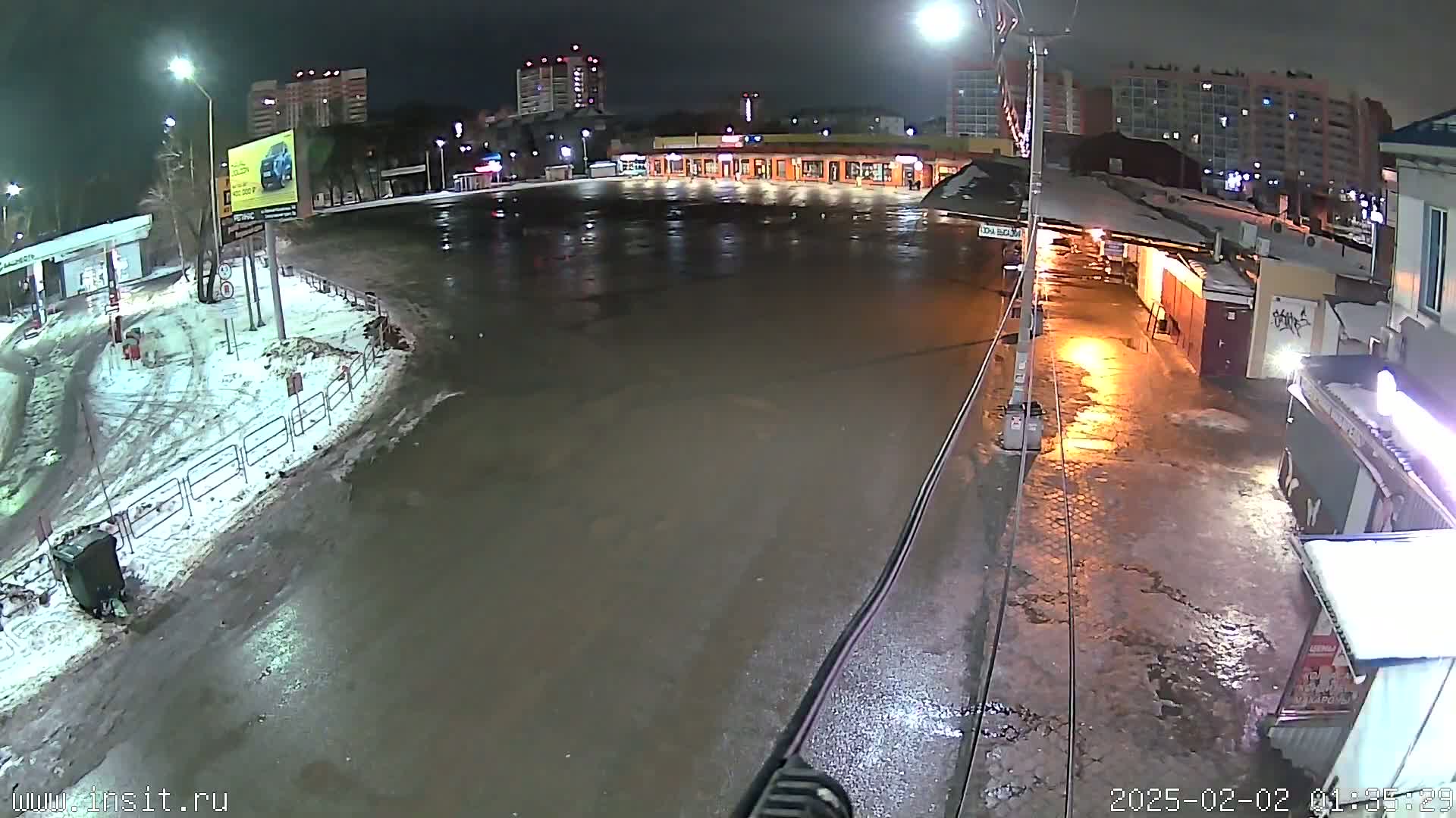 A wet, nighttime street scene shows a mostly empty plaza with several buildings in the background, some snow and ice on the ground, and streetlights illuminating the area.