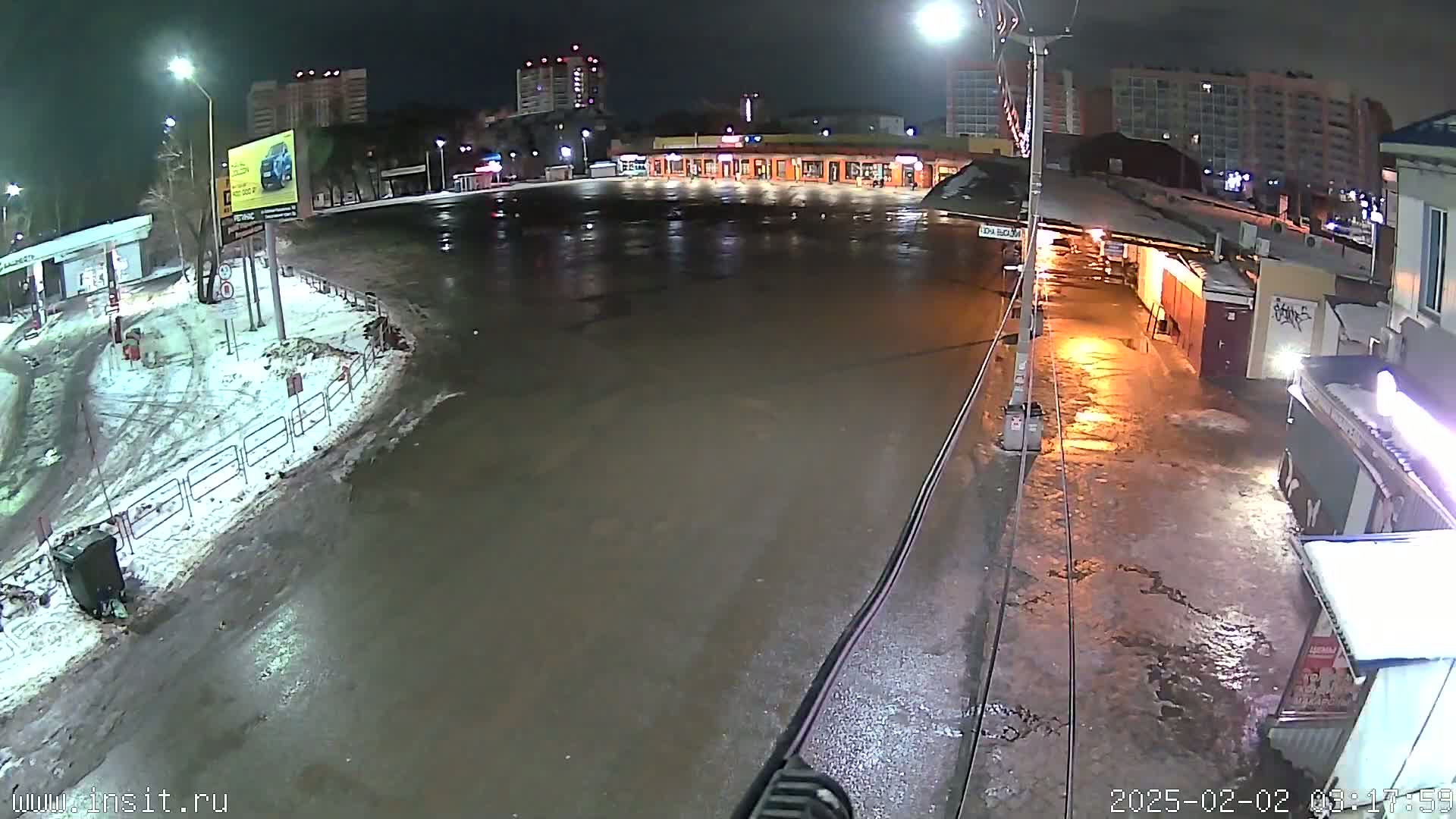 A wet, nighttime view of a mostly empty parking lot and surrounding buildings with some snow on the ground.