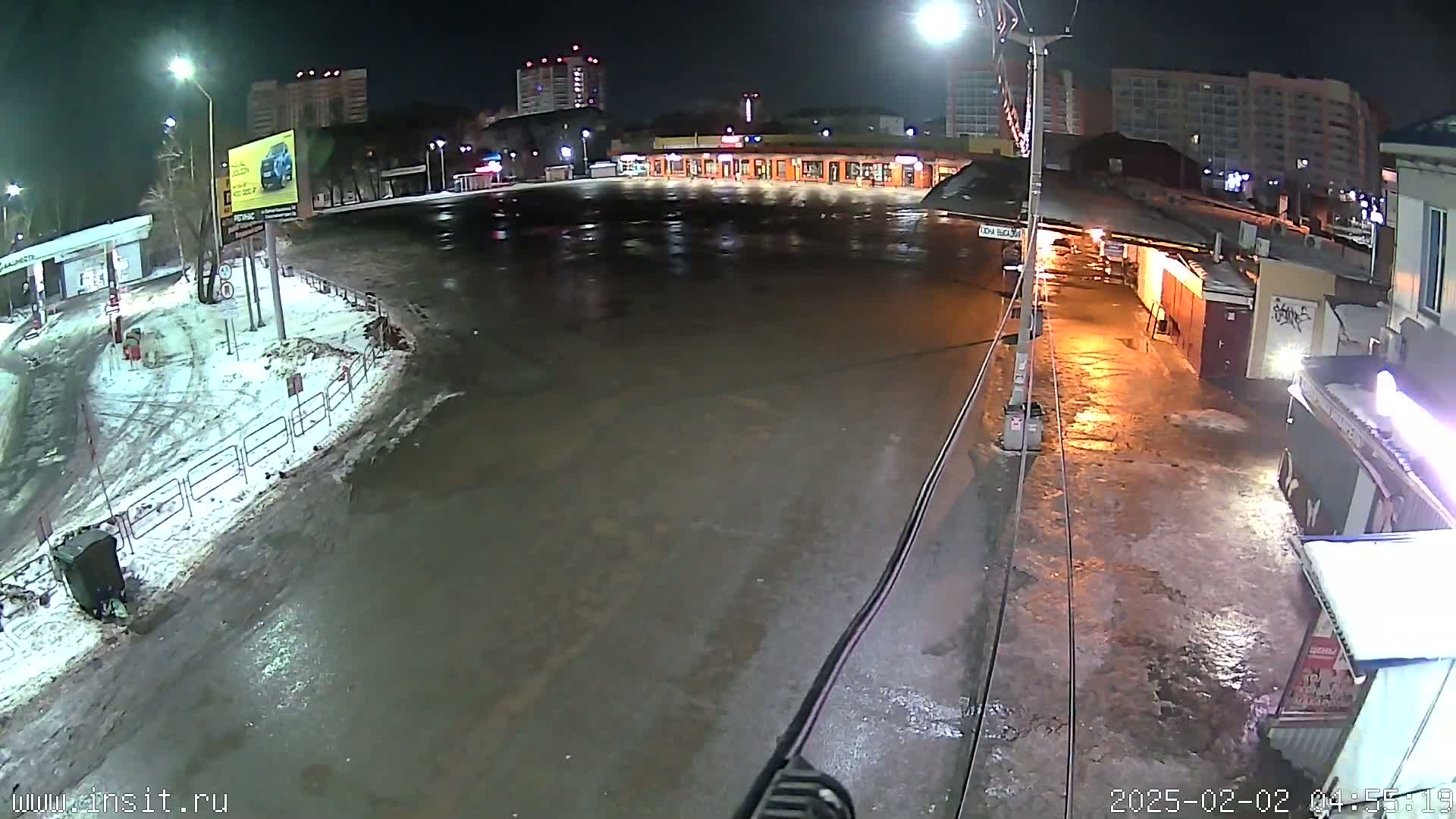 A wet, nighttime parking lot is seen with snow on the ground and buildings in the background under streetlights.