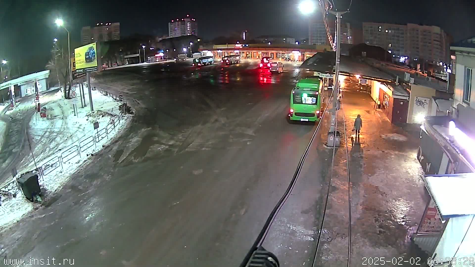 A nighttime, snowy scene shows a bus station with several buses, a person walking, and wet pavement.