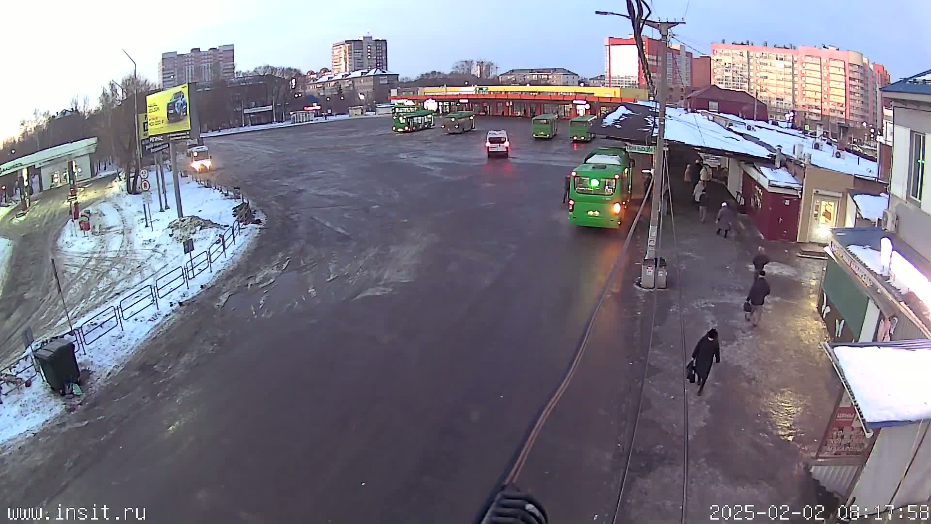 A bus stop with several buses and a few cars and pedestrians, in a snowy and icy area.