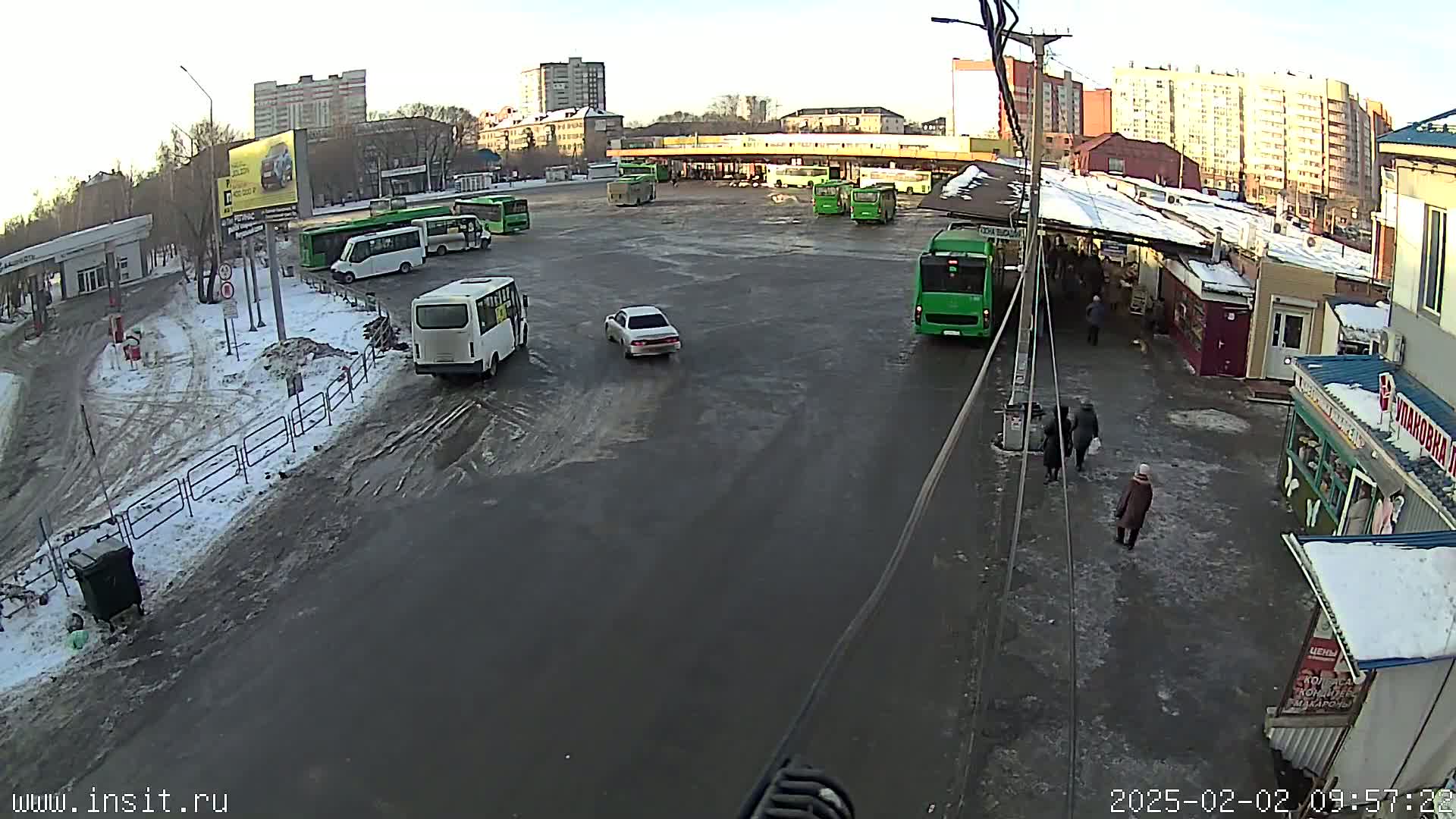 A mostly empty bus station with several buses and a few cars, on a partly snowy and slushy day.
