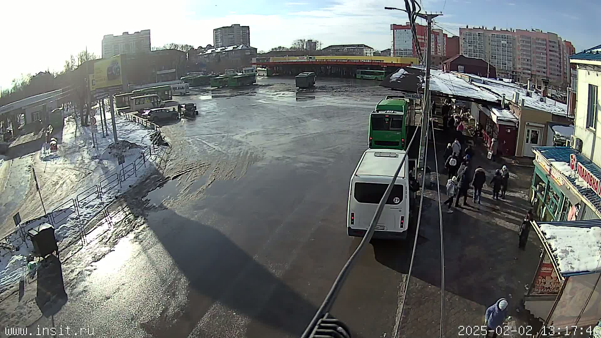 A bus station with several buses and people, partially covered in snow and ice, on a sunny day.