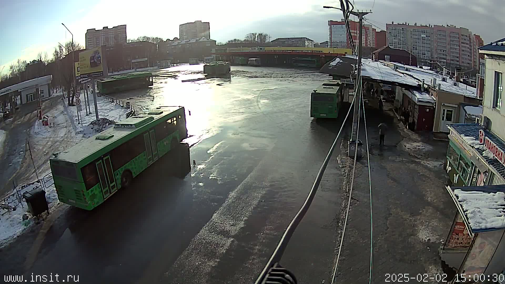 A mostly flooded bus depot is surrounded by apartment buildings under a partly sunny sky with some snow on the ground.
