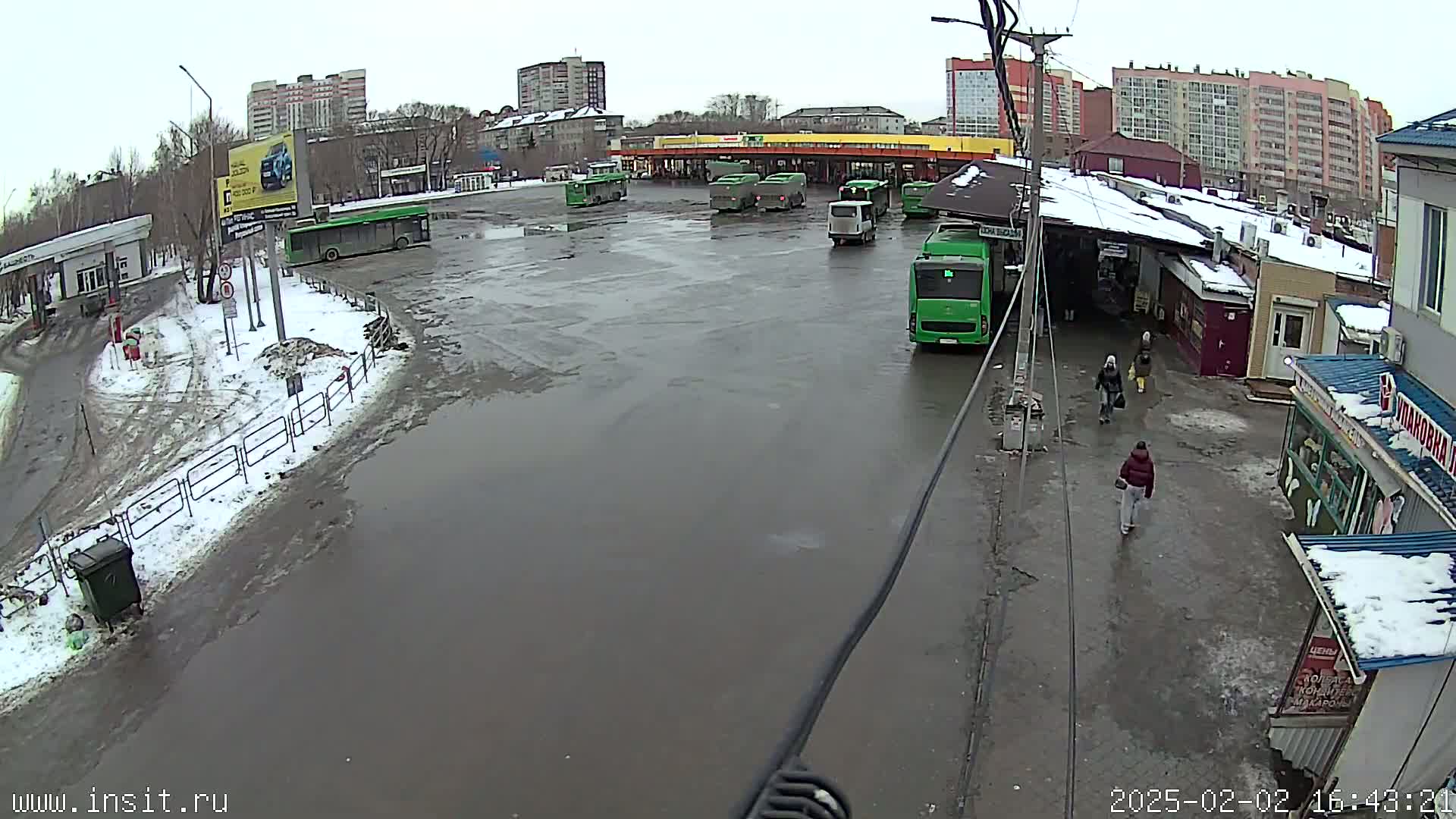 A bus depot with several buses parked in a large, wet lot, surrounded by buildings in snowy, overcast weather.