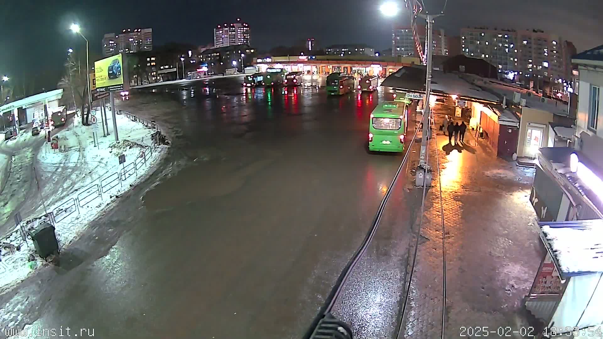 A wet, nighttime bus station with several buses parked and a few people standing near a building.