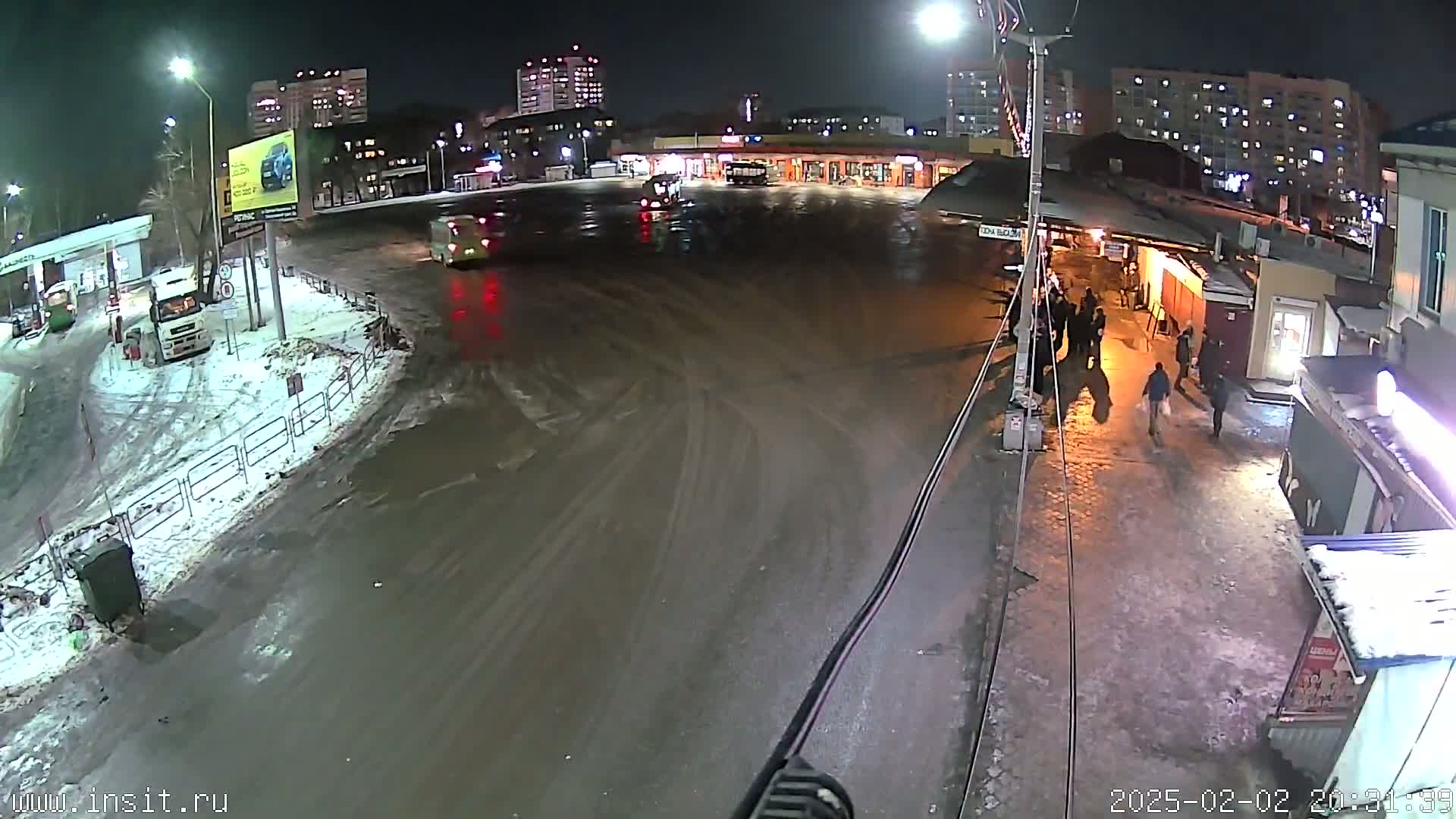 A nighttime, snowy scene shows a wet, tire-tracked roadway in front of a bus stop and several buildings, with a few vehicles and pedestrians present.