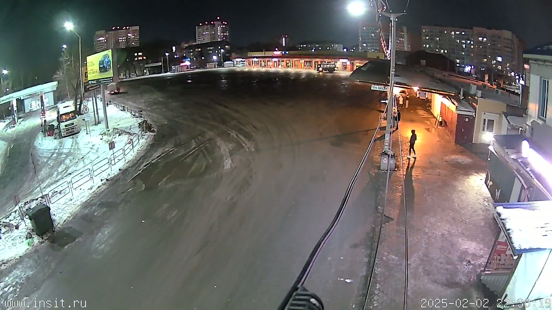 A nighttime, snowy scene shows a mostly empty bus station and surrounding area with a person walking near some buildings.