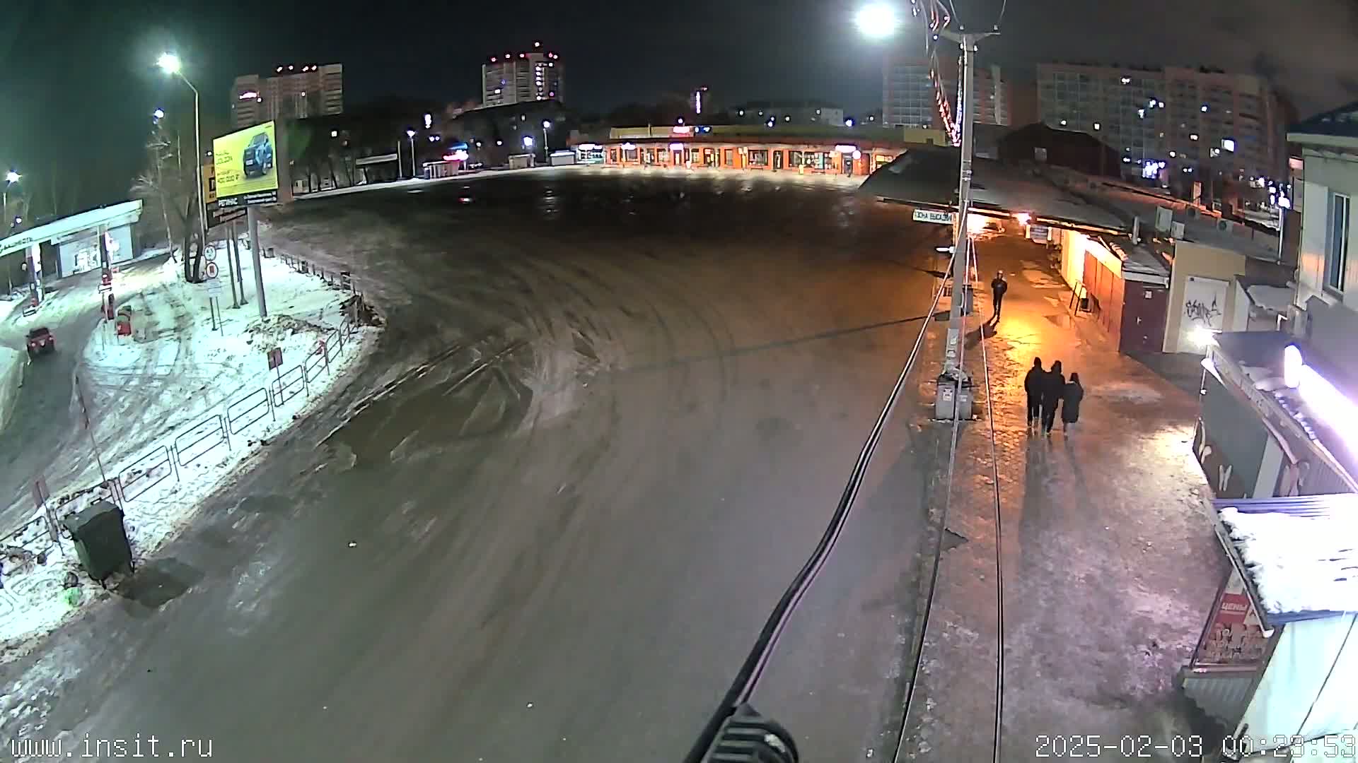 A nighttime, snow-covered and icy parking lot with several people walking and a few buildings visible in the background.