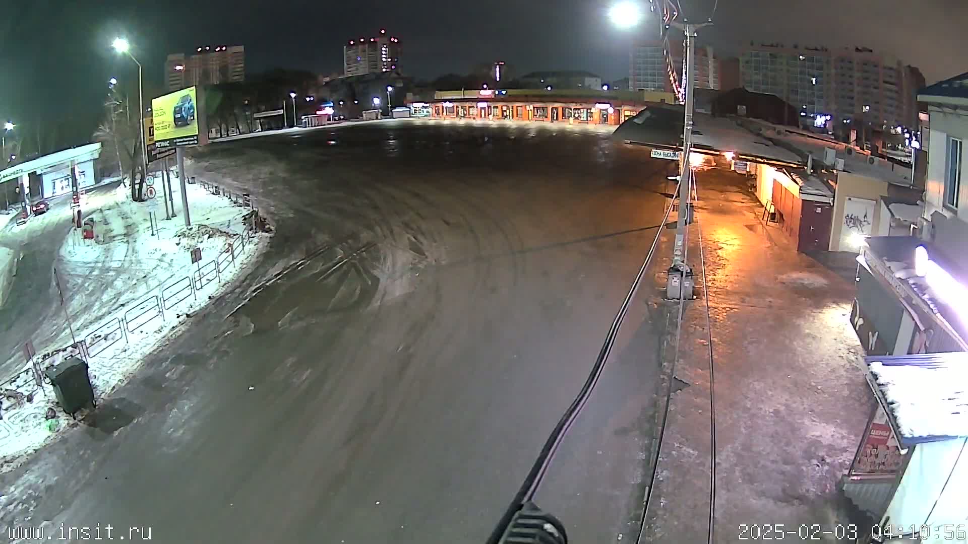 A snow-covered, mostly empty parking lot at night, illuminated by streetlights, with some buildings and a gas station in the background.