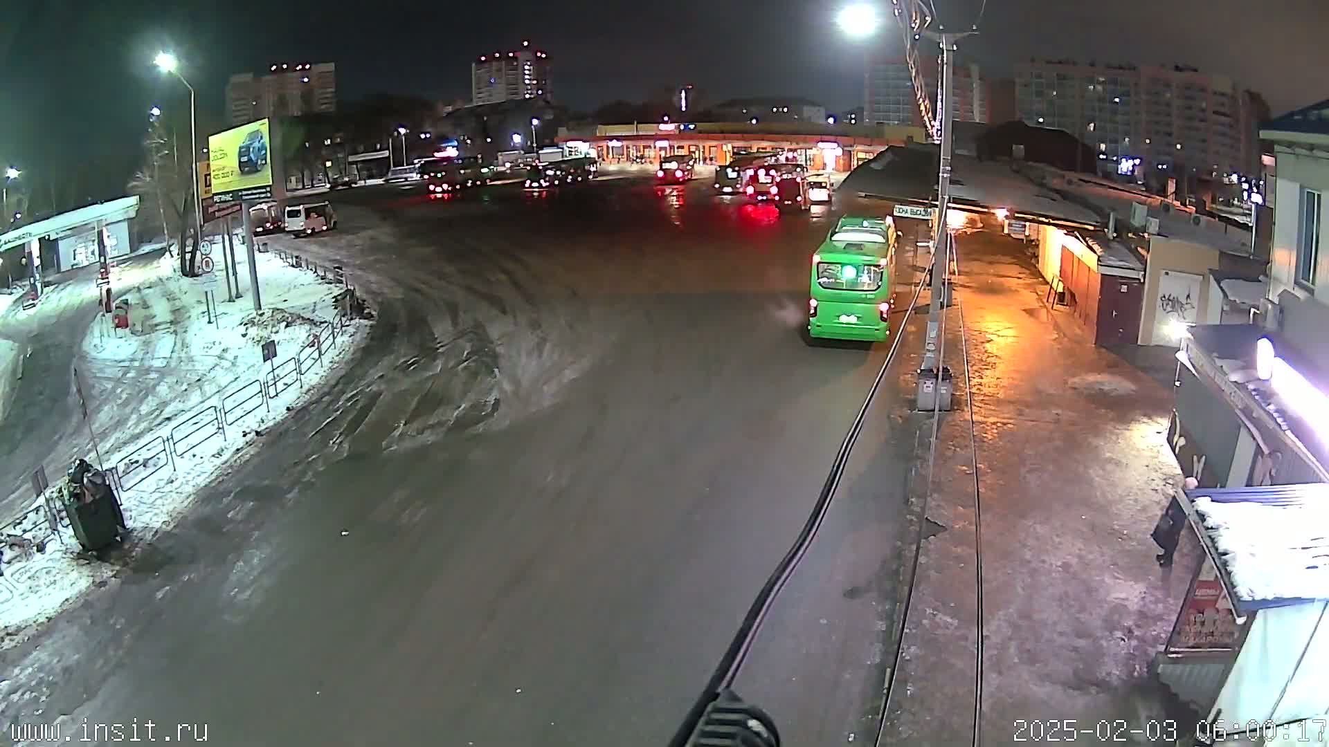 A nighttime, snowy scene shows a green bus at a bus stop with several other vehicles and buildings in the background.