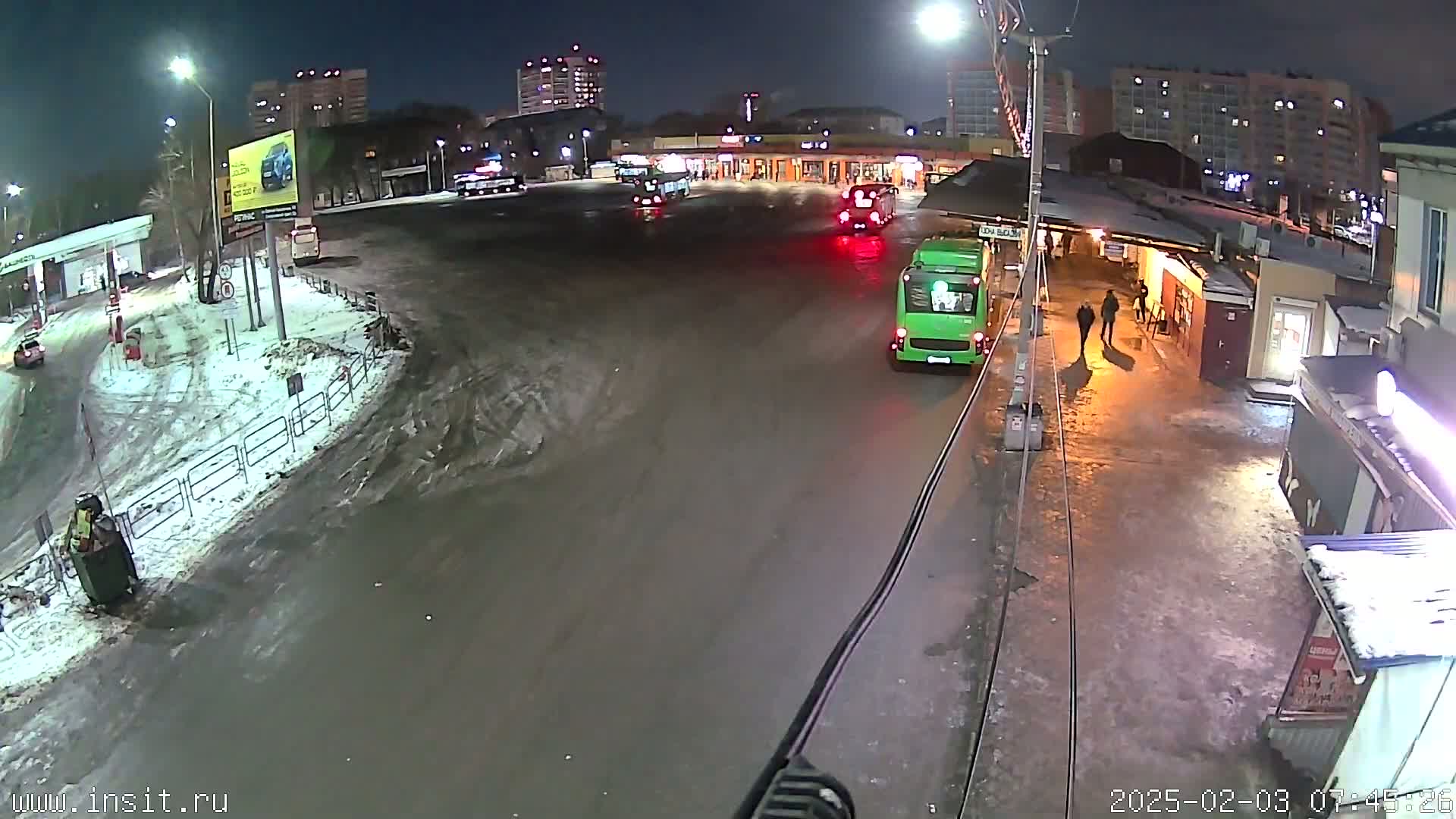 A nighttime scene shows several buses at a bus stop in a snowy, wet area with people walking nearby and apartment buildings in the background.