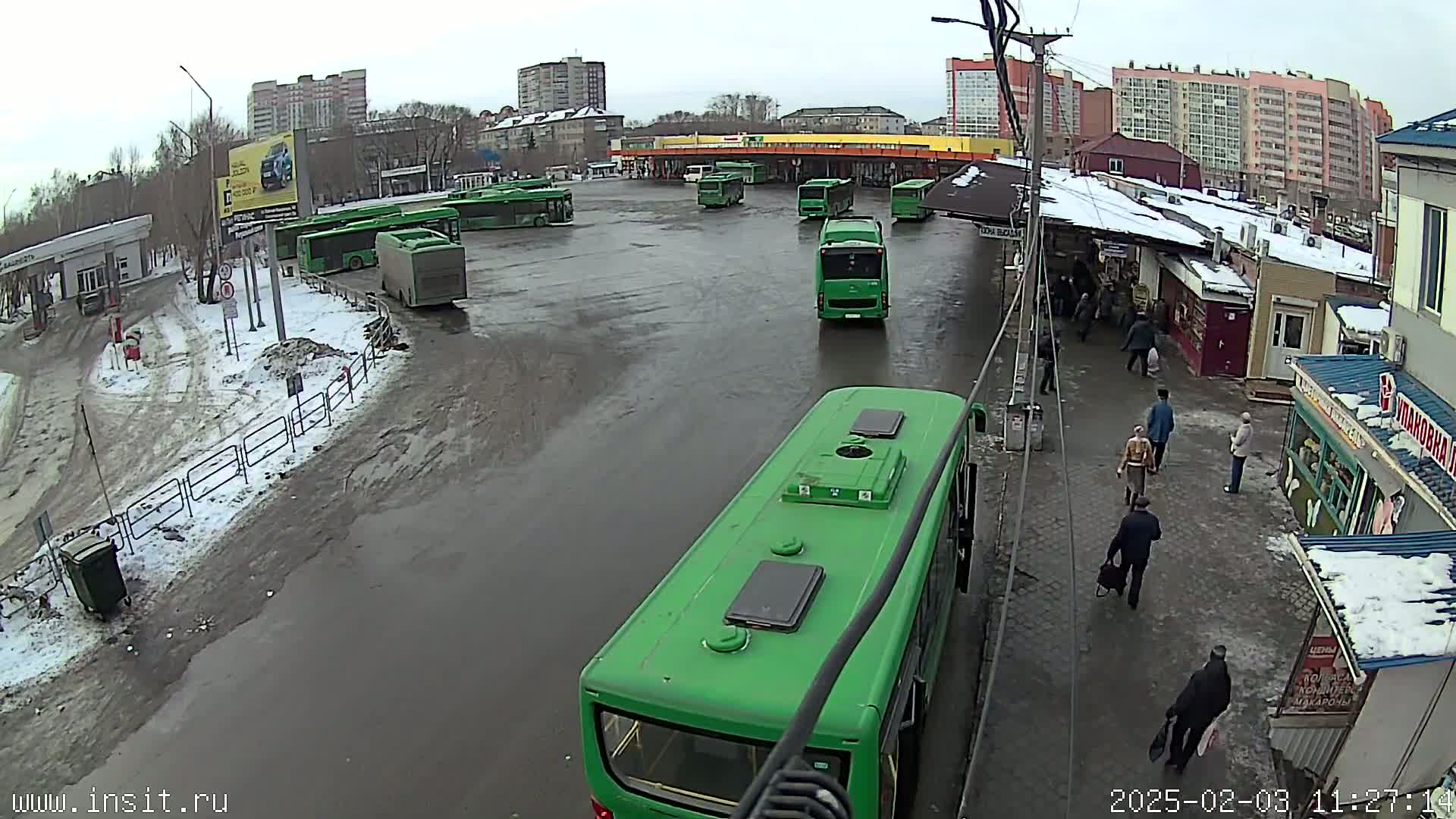A bus station with numerous green buses parked in a wet, partially snow-covered lot, surrounded by buildings and several pedestrians.