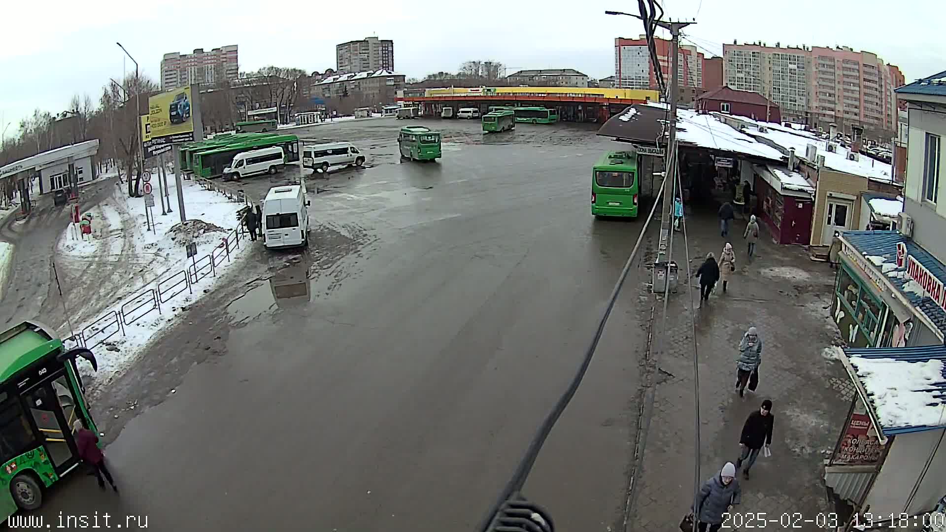A slushy, wet bus station with several green buses and people walking around, surrounded by residential buildings under an overcast sky.