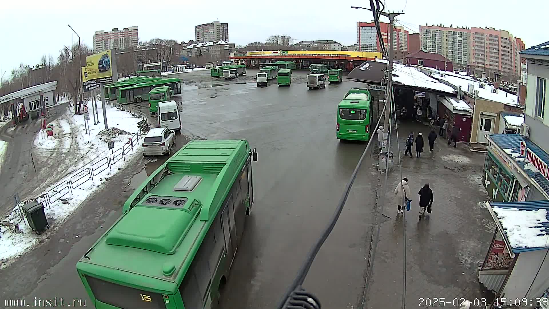A bus depot with numerous green buses and several cars parked in a wet, snowy area, with people walking around nearby buildings.
