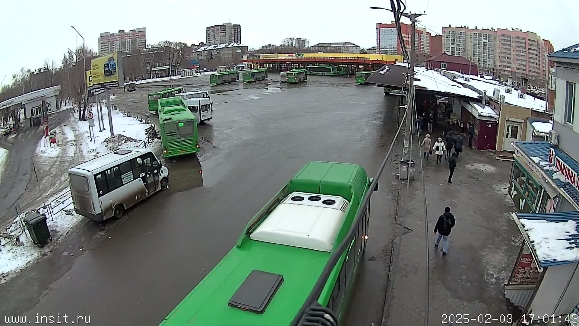 A bus depot with numerous buses parked in a wet, snowy area, with several people walking around nearby buildings.