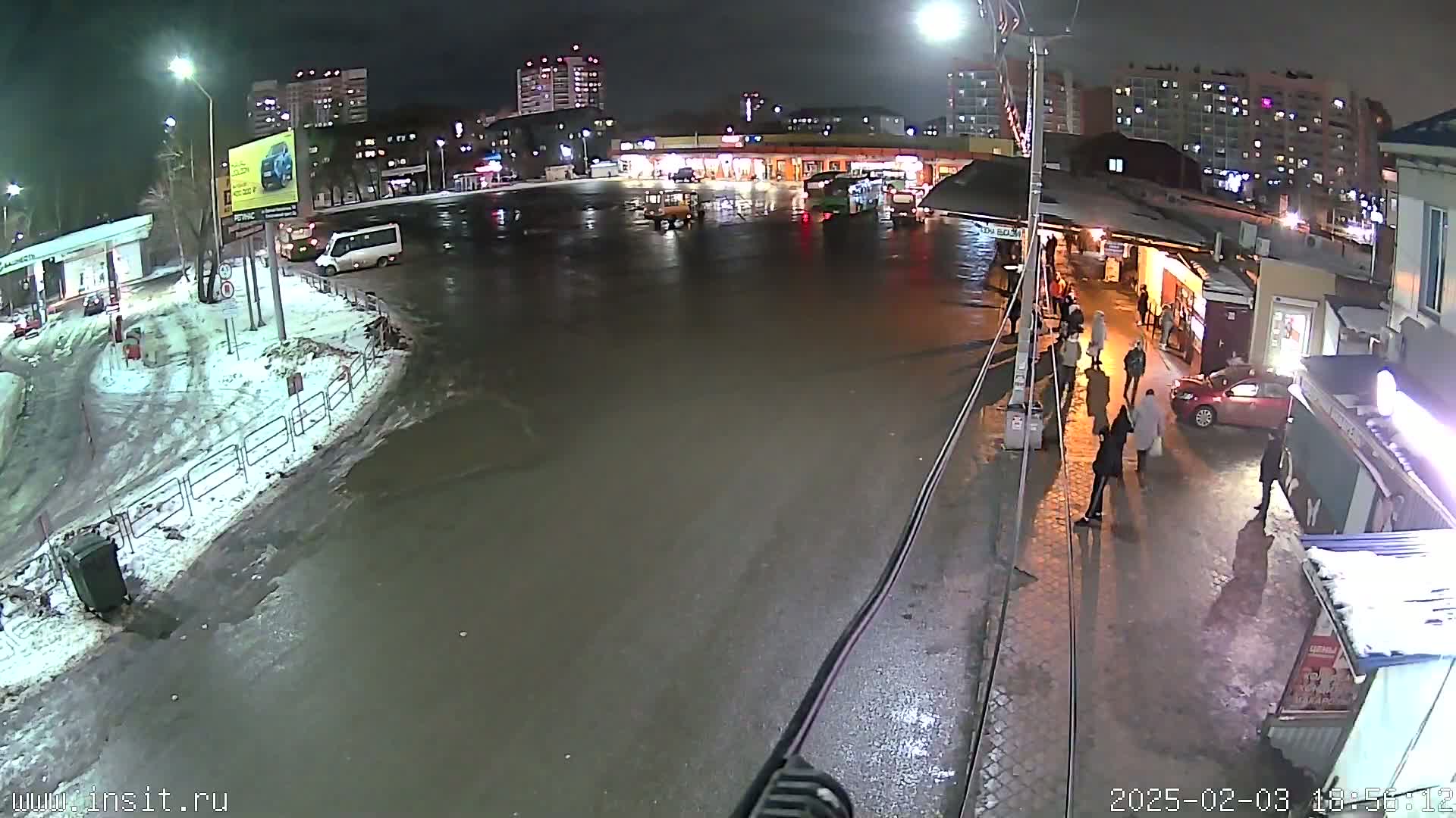 A wet, nighttime scene shows a bus stop with several people standing near buildings, vehicles parked in a lot, and some snow on the ground.